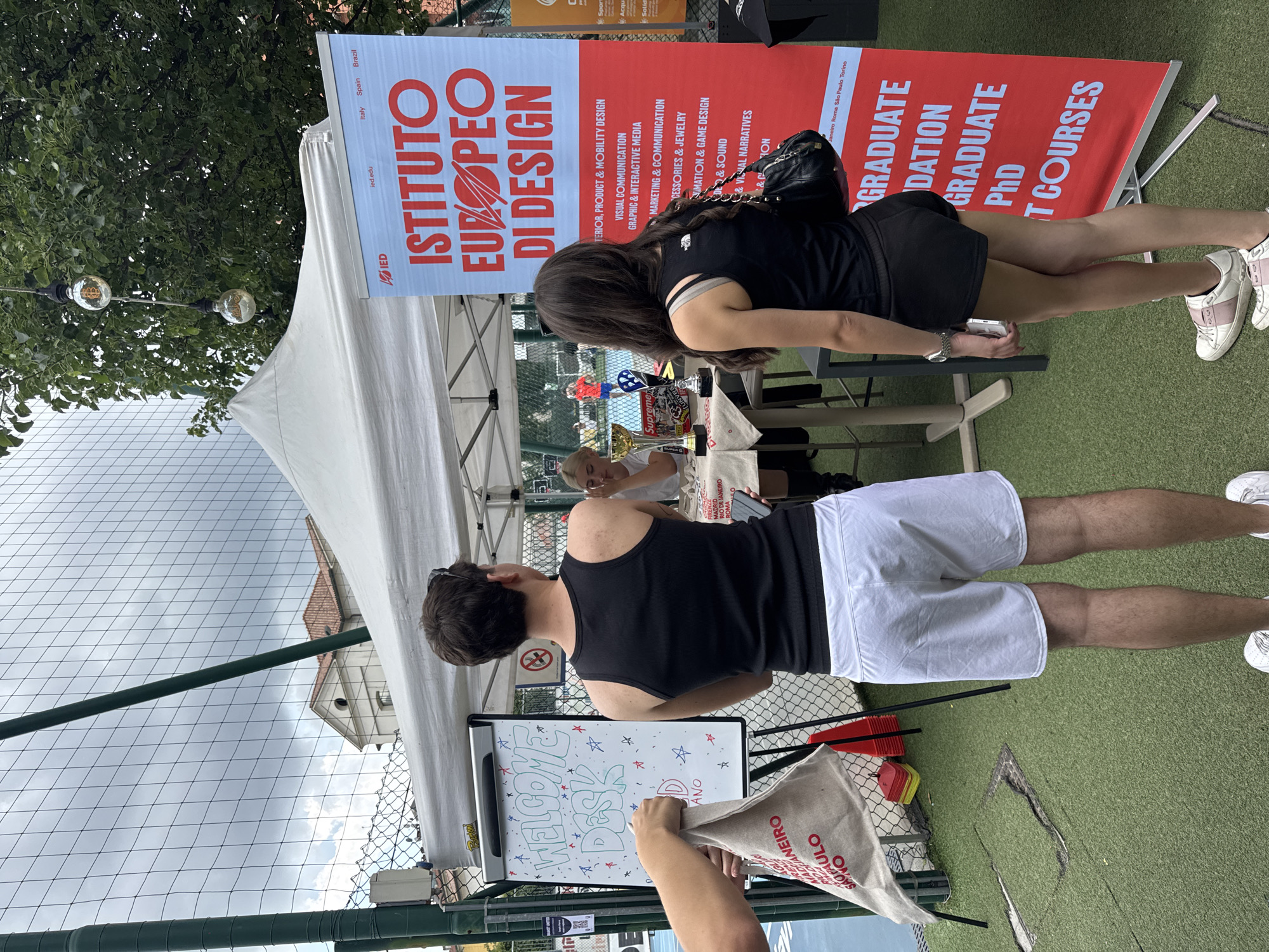 This image captures an outdoor promotional event for Istituto Europeo di Design (IED) in Milan, Italy. The scene is set on artificial green turf, possibly within a sports complex, enclosed by a green chain-link fence.
In the foreground, a male and a female stand with their backs mostly to the camera, facing a white canopy tent and a large red banner. The male, on the left, wears a black tank top and white shorts, with white sneakers. The female, on the right, wears a black tank top with a "The North Face" logo on the strap, black shorts, and white and pink sneakers. She carries a black handbag over her right shoulder and holds a white smartphone in her left hand.
The red IED banner, partially visible on the right, prominently displays "ISTITUTO EUROPEO DI DESIGN" in white lettering and lists various design disciplines such as "INTERIOR, PRODUCT & MOBILITY DESIGN," "VISUAL COMMUNICATION," and "GRAPHIC & INTERACTIVE MEDIA," among others. At the top, "ied.edu" and "Italy Spain Brazil" are visible, with "Janeiro Roma São Paulo Torino" listed at the bottom, indicating IED's international presence.
To the left, within the booth area, a "WELCOME DESK" is visible on an easel-mounted whiteboard, decorated with stars. A blonde female in a white top is seated at a table behind this desk, partially obscured. A gold-colored trophy and another smaller award are visible on the table in front of her. A person's hand, entering from the left, holds a light-colored cloth bag with red text listing cities like "JANEIRO" and "SAO PAULO."
Overhead, two decorative light bulbs, one containing a Bitcoin symbol, hang from a tree branch. Red and yellow traffic cones are stacked near the fence behind the "Welcome Desk," next to a "no smoking" sign. In the background, beyond the fence, a building with a terracotta tiled roof is visible against a cloudy sky, and faint figures are seen on a blue sports court.