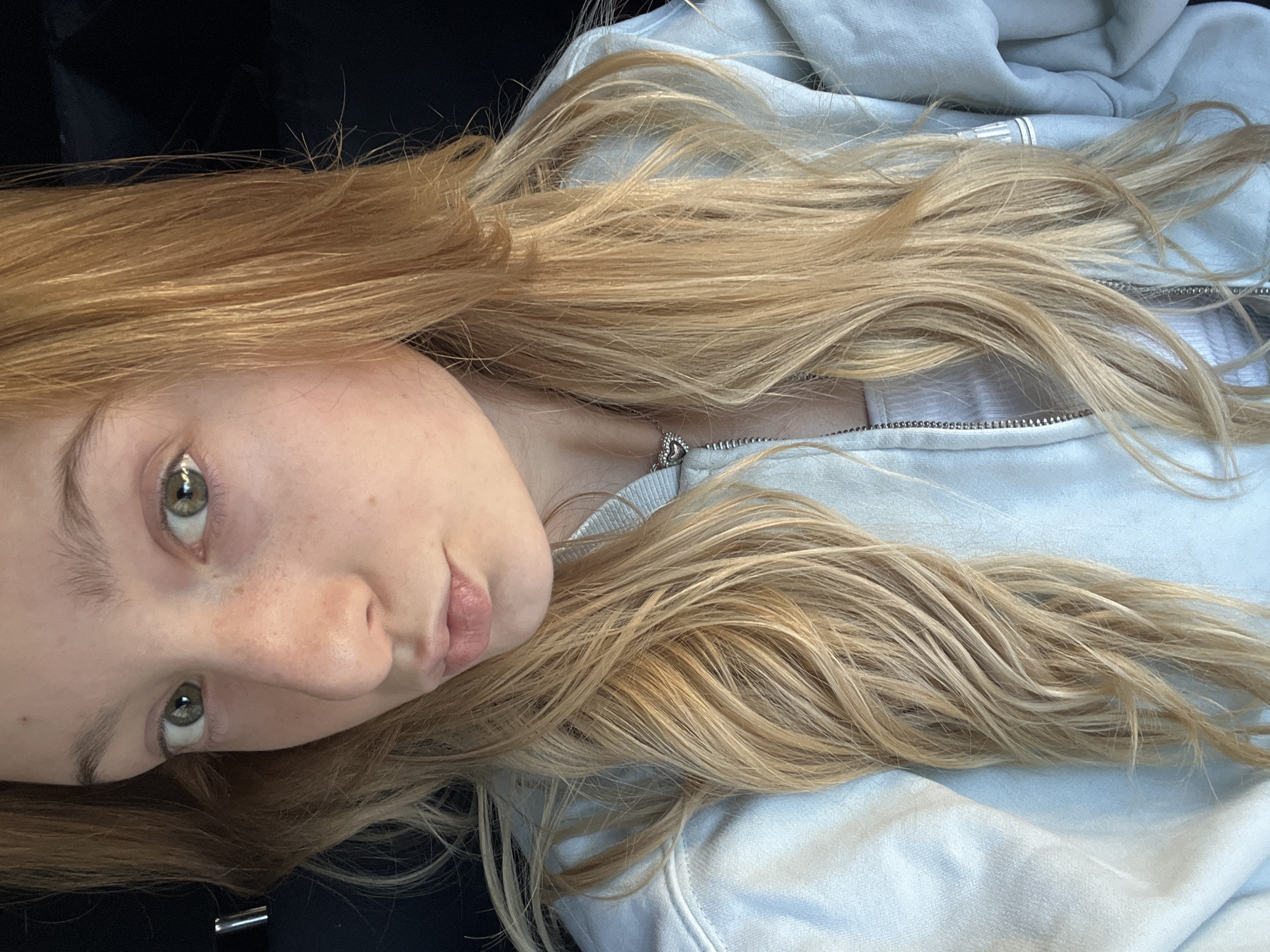 A young female individual is depicted in a close-up, chest-up portrait. She possesses long, wavy blonde hair with lighter highlights, which cascades over her shoulders and partially covers her light blue, zippered hoodie. Her facial features include light green eyes, a small scattering of freckles on her nose and cheeks, and her lips are slightly parted in a pout. She wears a plain white undershirt visible beneath the open zipper of the hoodie, and a silver chain with a small heart-shaped pendant is visible around her neck. The background is dark and indistinct, suggesting an interior setting such as a vehicle or room with minimal ambient light. No specific geographic identifiers are present in the image to determine a location within the city of Varna, Bulgaria, or any other specific city.