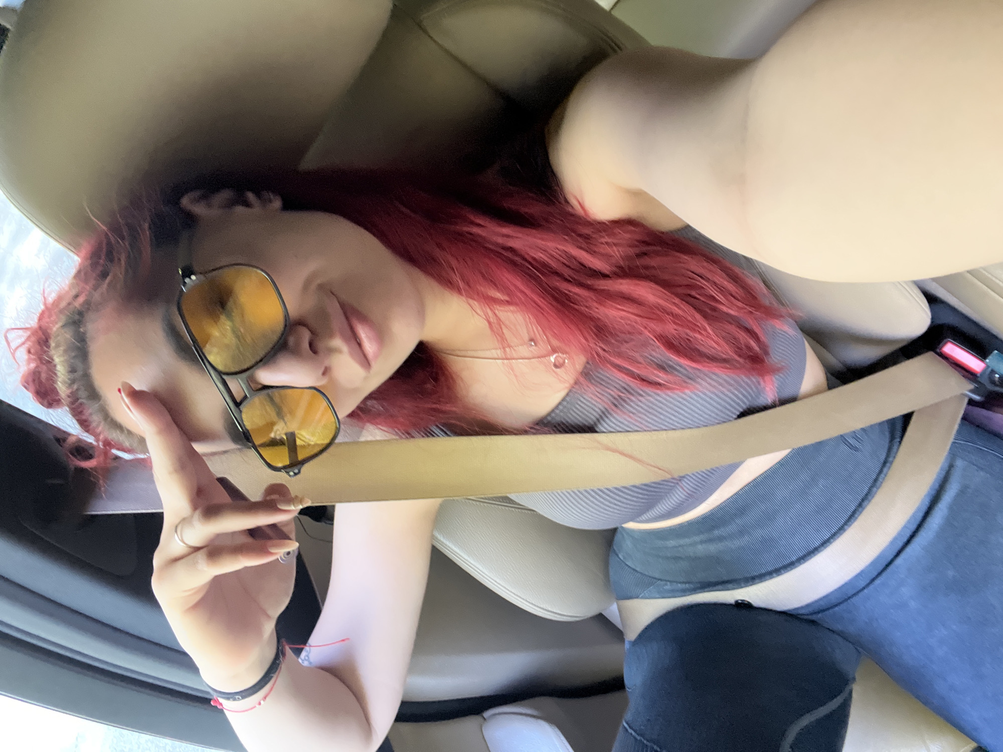 A selfie captures a woman inside a car, likely in the passenger seat, during daytime. The woman has long, vibrant red hair and is wearing yellow-tinted aviator-style sunglasses. Her lips are slightly parted. She is dressed in a grey ribbed crop top and dark blue or grey leggings. A light brown seatbelt stretches diagonally across her chest and lap, indicating she is belted in. Her left hand, adorned with a silver ring on her index finger and what appears to be a bracelet or band on her wrist, is raised to her forehead in a casual pose. Her fingernails are long and light-colored.
The interior of the car features light brown or tan leather seats, and parts of the car's door or dashboard are visible. Bright natural light streams in from outside the vehicle, suggesting a sunny day. No specific outdoor landmarks or text are visible to identify the exact location within Vrata, Bulgaria, as provided in the context. The overall mood conveyed is casual and relaxed, fitting for a self-portrait taken while traveling.