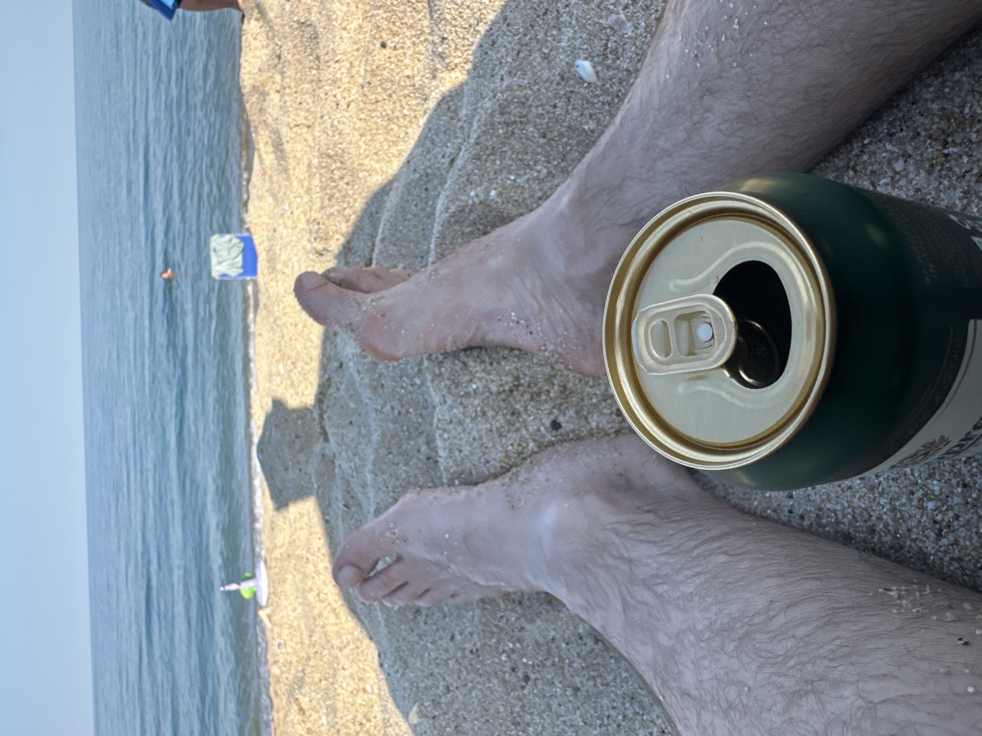 The image shows a close-up view of a person's legs and feet resting on a sandy beach, with the ocean in the background. A green and gold beverage can is placed next to the person's legs. The sand appears wet in some areas, and small shells or debris are scattered across it. The ocean water is calm and a light blue, with subtle ripples. In the distance, a person can be seen swimming in the ocean, and a small, colorful object, possibly a toy, is floating near the shoreline. The lighting suggests it is daytime, likely sunny, given the bright sand and the shadows cast. The overall mood is relaxed and casual, typical of a beach setting. No visible text is clearly legible on the beverage can or any other element in the image.