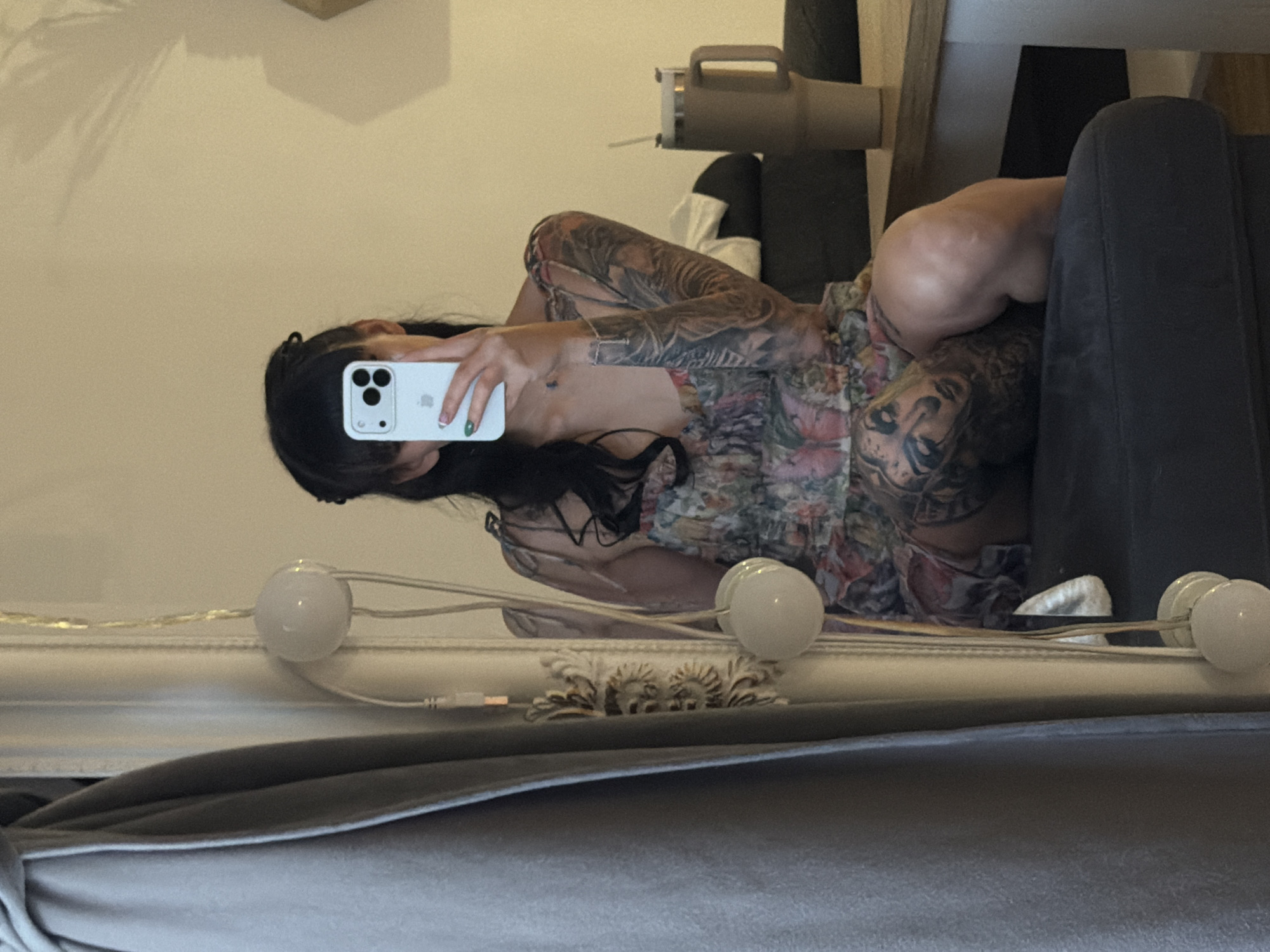 The image captures a selfie reflection in a mirror, depicting a woman lying down indoors. The woman, with long dark hair, is visible from the chest up, holding a white smartphone to take the picture or view its screen. She has extensive tattoos covering both arms and parts of her torso, and is wearing a floral-patterned top or dress. Her fingernails are painted, with one visible nail sporting green polish.
She appears to be reclining on a dark gray or black couch or armchair, which occupies the right side of the frame. In the background, behind her head, a light-colored object with a handle, possibly a humidifier or a stylized lamp, is visible. The wall reflecting behind her is a light, neutral color, with faint shadows of plant leaves or window frames cast upon it.
Along the bottom of the reflected image, a string of white spherical lights is draped, with a visible white cord and a USB-A connector. Below the string lights, ornate, decorative molding or trim is present. The overall setting is an indoor space, likely a living room or bedroom, with artificial lighting contributing to the ambience. There is no visible text in the image. Given the provided context, this scene is located in Plovdiv, Bulgaria.