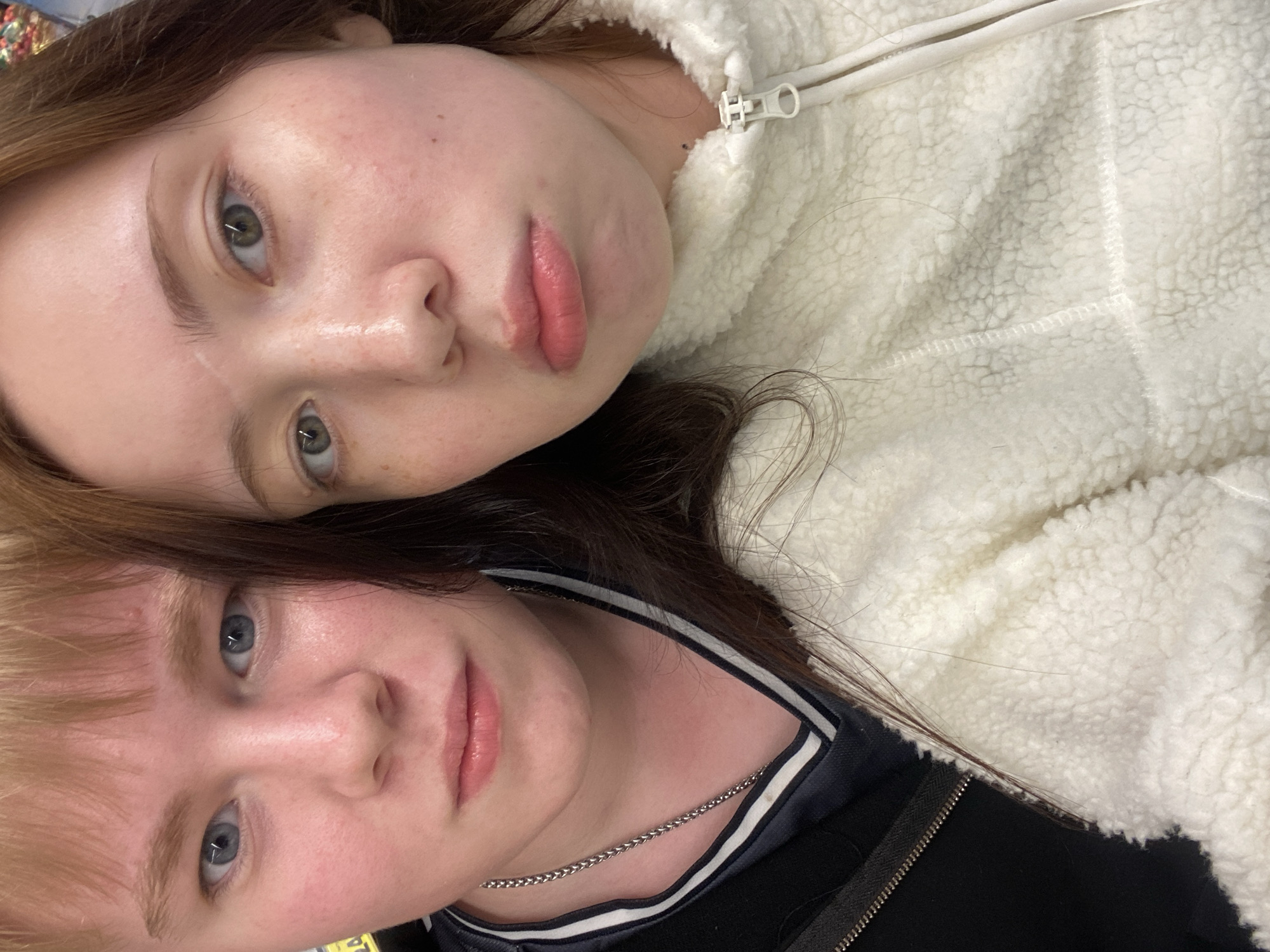 Two individuals are closely positioned, face-to-face. The individual at the top of the image has light skin, green eyes, and auburn hair. They are wearing a white, fleece-like jacket with a zipper. The individual at the bottom of the image also has light skin, blue eyes, and blonde hair. They are wearing a black jacket with a white stripe near the collar, and a silver chain necklace is visible around their neck. The background is indistinct. The location is Unknown, Unknown.