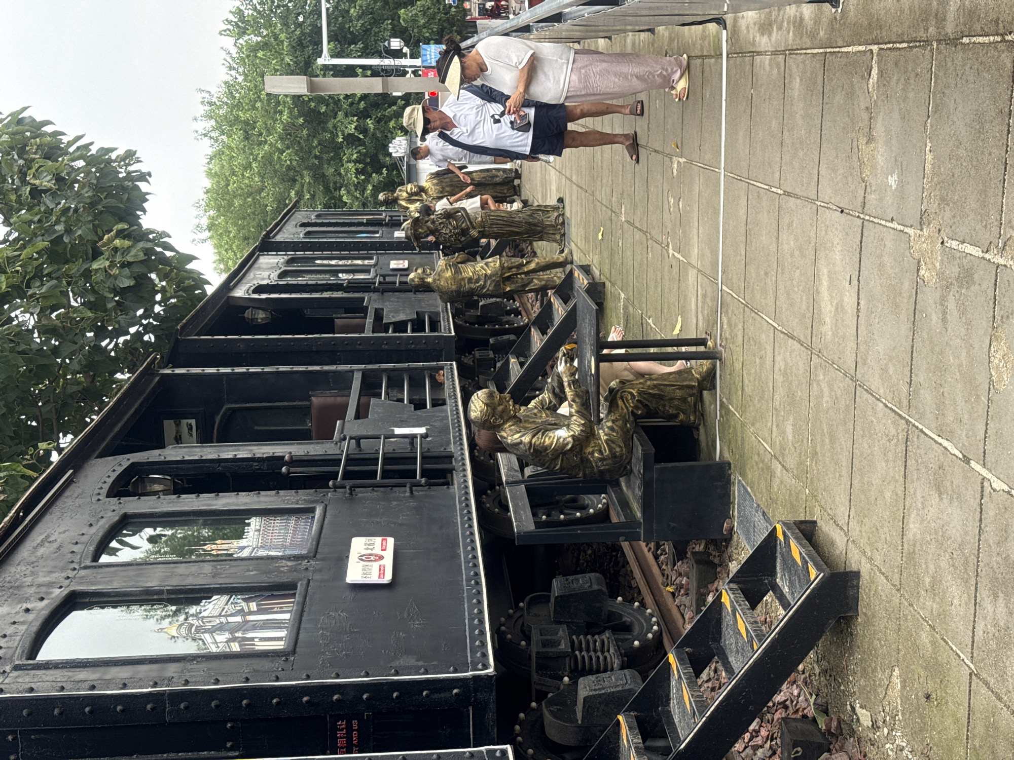 The image depicts a static outdoor scene featuring a vintage black train car on tracks, located at the "Binhai Road Historic Train Park" in Qinhuangdao, China. The train car is adorned with reflective decals on its windows, showcasing architectural facades. In front of the train, a row of bronze statues depicts individuals in historical attire, some engaged in various activities like sitting at a table or standing in conversation.
Several individuals are present in the scene. Two are standing on a tiled walkway in the foreground, observing something on a mobile phone. One is a man wearing a white polo shirt, dark shorts, and a straw hat. The other is a woman in a white shirt and a wide-brimmed hat, also looking at a phone. Further back, more people are visible, some interacting with the statues and others walking along the path. The environment includes lush green trees in the background, a cloudy sky, and the train tracks with loose gravel and ballast. The walkway is paved with large square tiles, some showing signs of wear. The scene appears to be a tourist attraction, with clear signage and the presentation of historical elements.