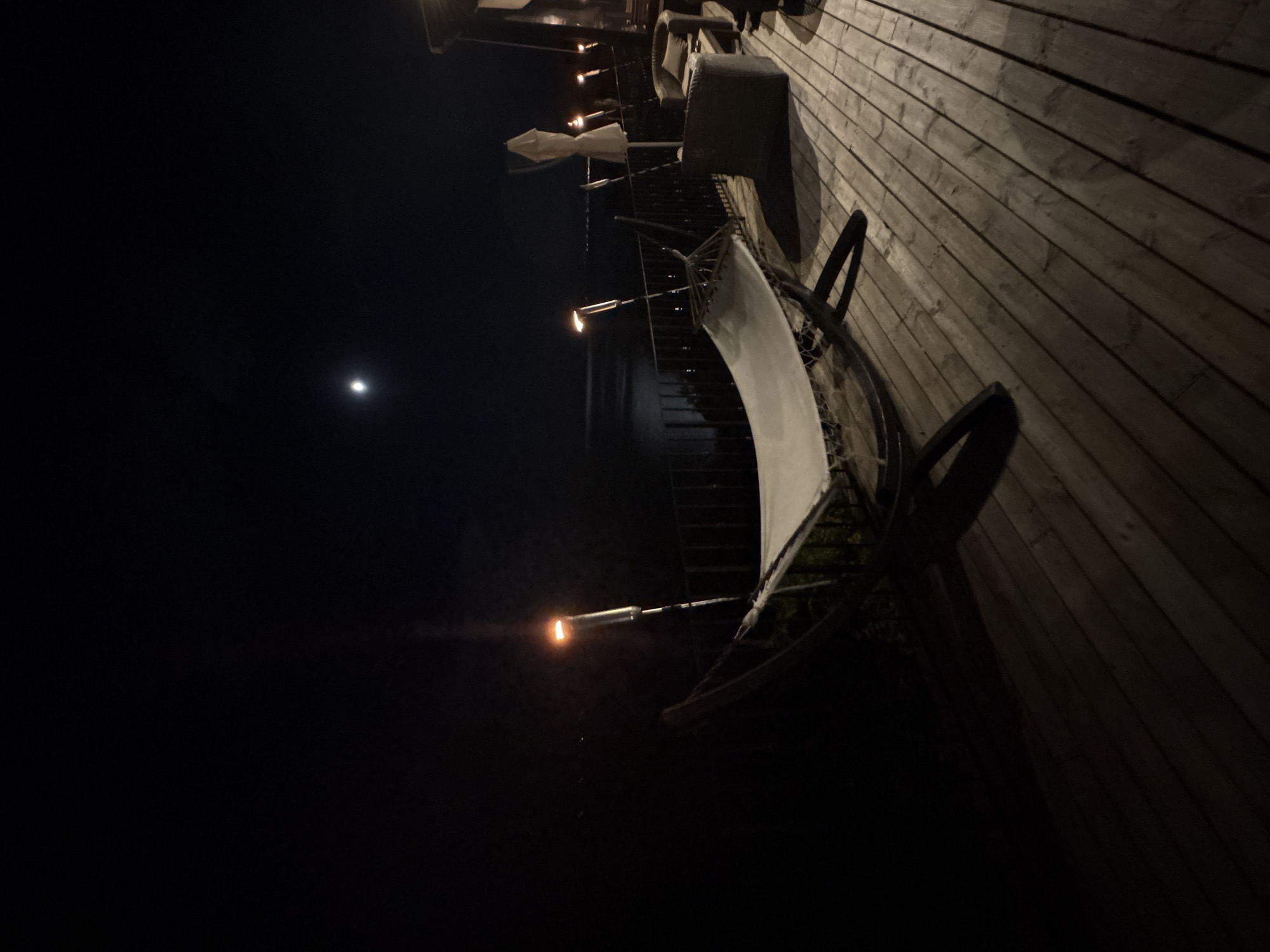 The image depicts an outdoor patio area at night, illuminated by several torch lights and what appears to be a full moon in the dark sky. The patio is constructed of wooden planks. Various objects are visible on the patio, including what looks like patio furniture such as chairs with cushions, and a white canvas stretched over a frame, possibly a shade or canopy. Several torch lights are mounted on railings, casting a warm glow. The scene suggests a relaxing evening outdoors. The presence of the moon and the torches indicate it is nighttime. The overall mood is serene and tranquil, with the darkness of the night contrasting with the warm light sources. There is no visible text in the image.