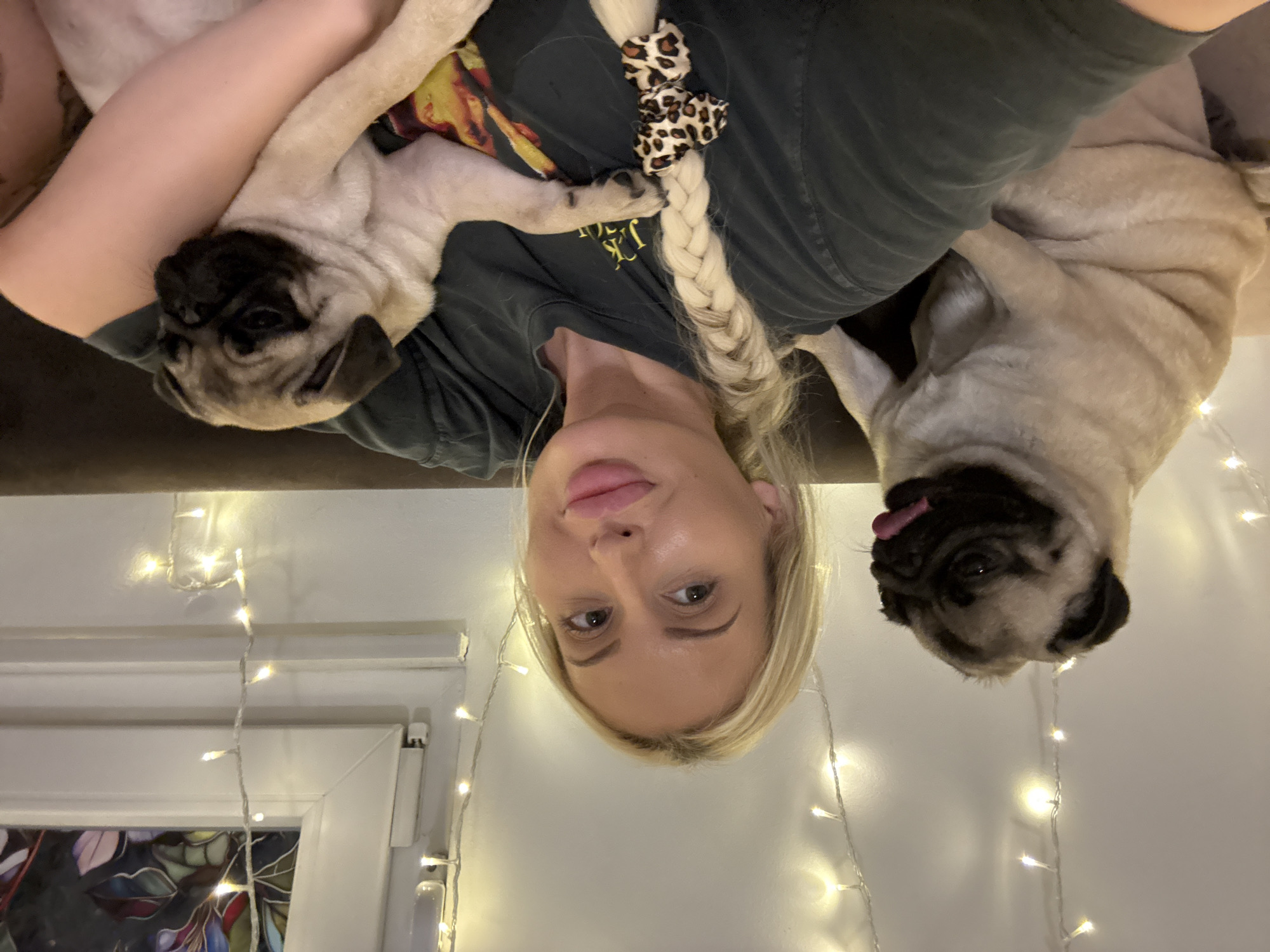 A blonde woman with full lips and full eyebrows, wearing a dark t-shirt, sits on a brown couch, holding a fawn-colored pug puppy. Her hair is styled in a single braid that falls over her shoulder and is adorned with a leopard-print hair tie. To her left, another fawn-colored pug sits upright with its tongue slightly out. The background is illuminated by a string of fairy lights draped along the wall. A window with decorative glass panes is visible behind the woman.