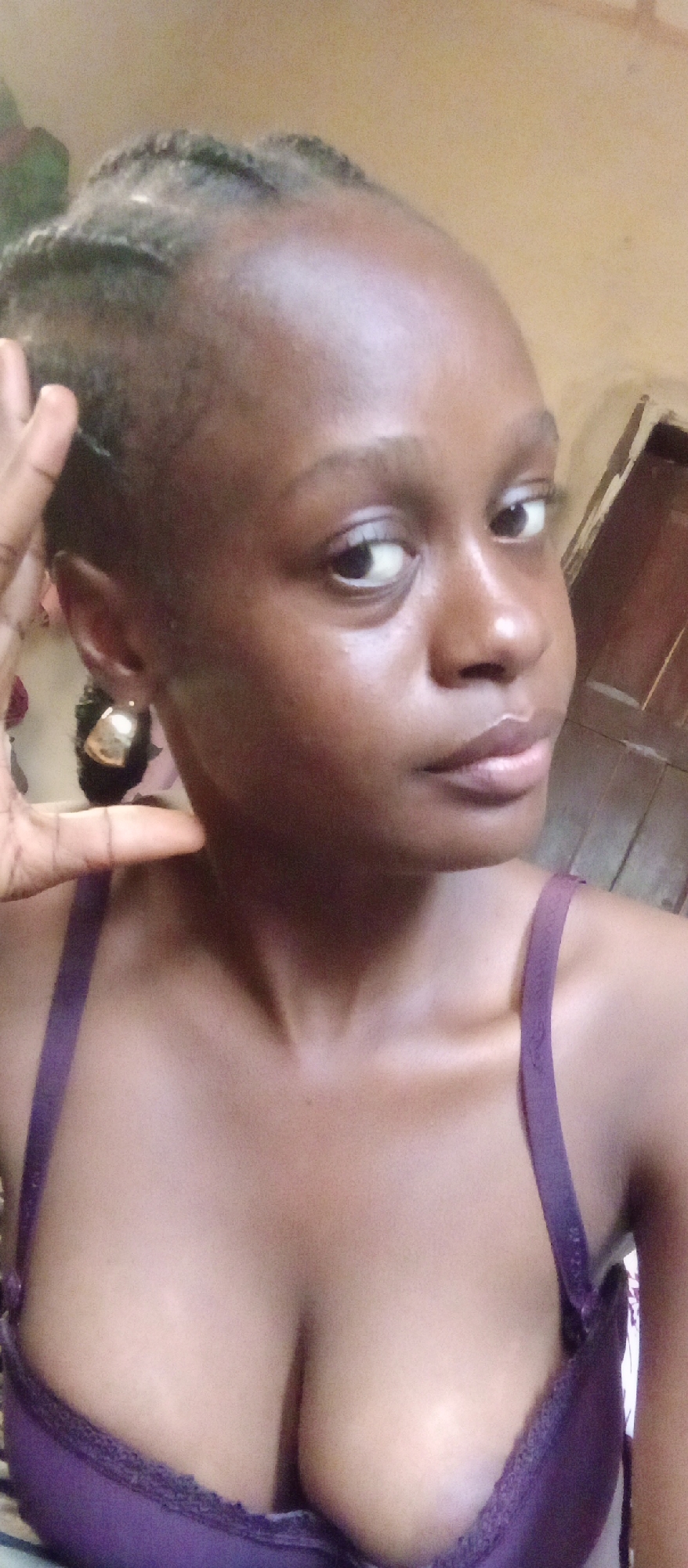 A medium close-up photograph shows a young Black woman with dark braided hair in Akwanga, Nigeria. She is wearing a dark purple bra and has gold hoop earrings. She is looking toward the camera with a neutral expression. Her right hand is raised to her face. The background is a slightly blurry, light beige wall and a dark brown door.