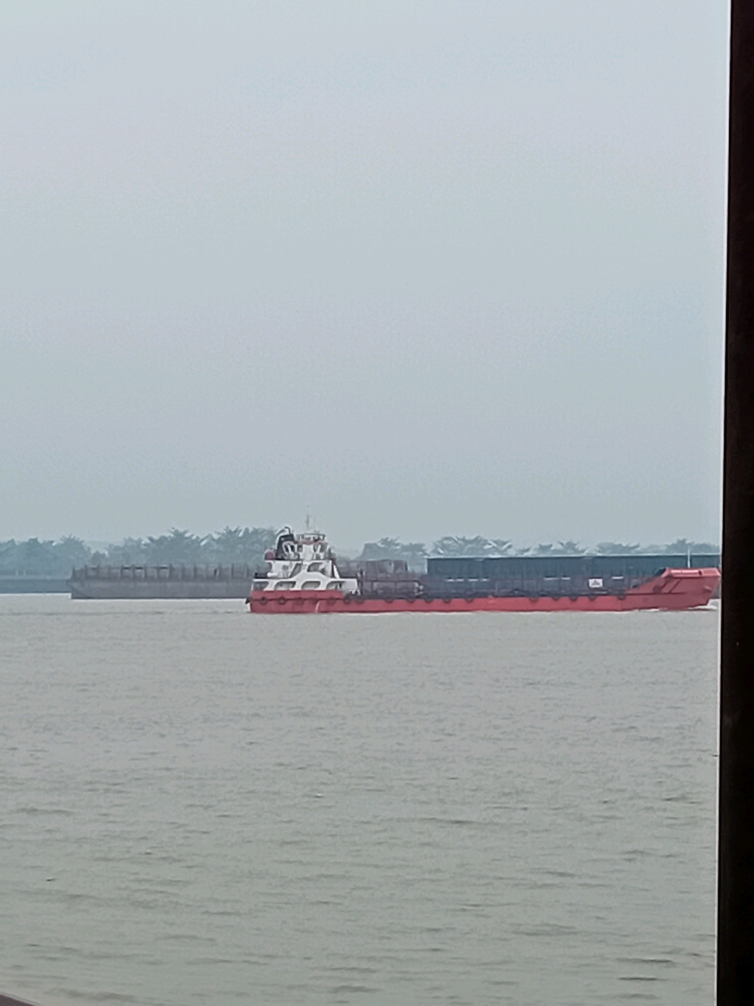 A cargo barge, painted red and white, with a dark cargo cover, navigates a wide, calm body of water, likely a river. In the background, a line of trees, possibly oil palm, marks the distant shore. Another, larger barge, mostly gray, is visible further in the distance to the left. The sky is overcast and hazy. The scene is set in Sungai Meriam, Indonesia, a region characterized by its extensive river systems and agricultural landscapes.