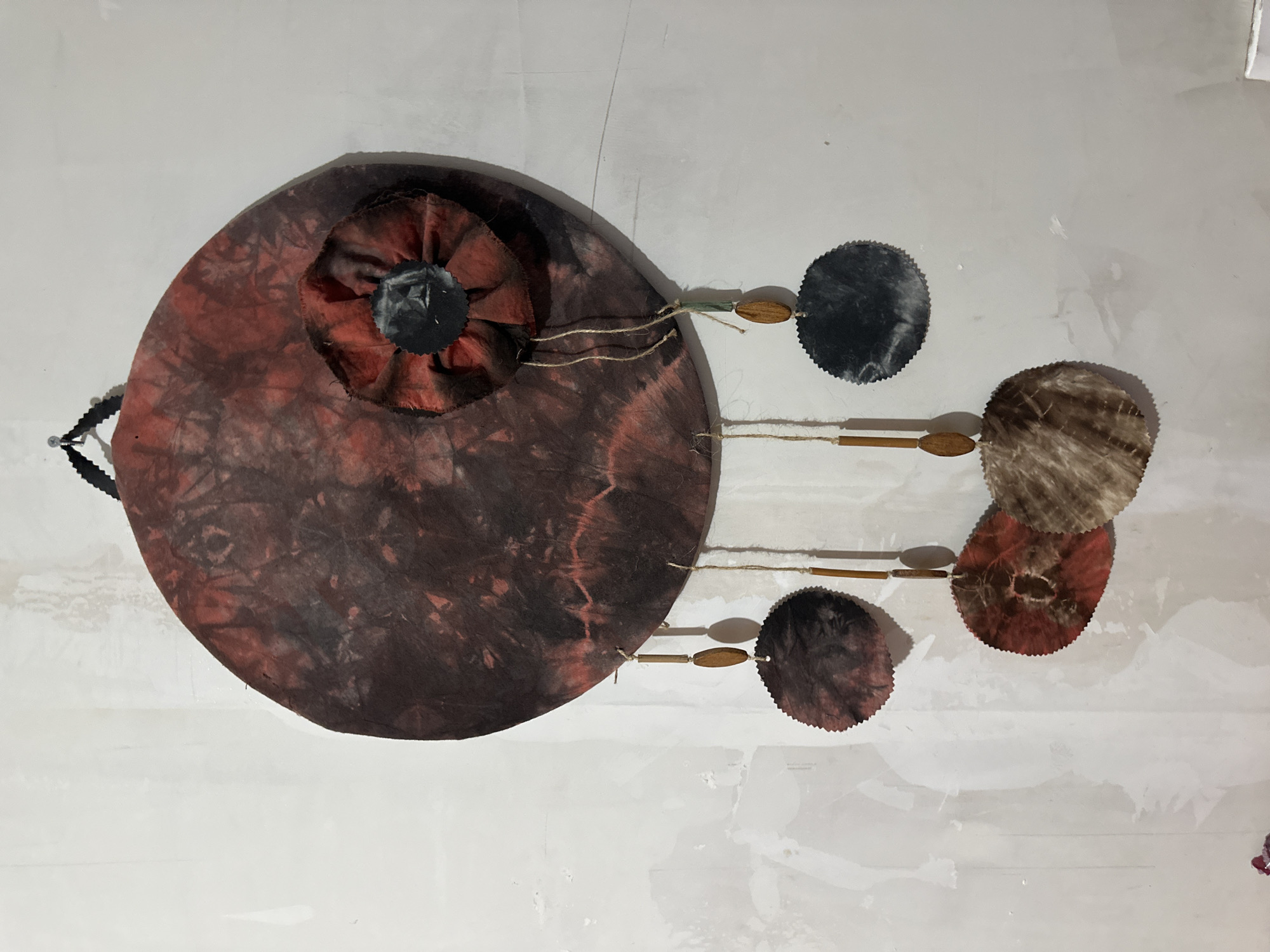 An overhead shot depicts a decorative wall hanging against a white wall, possibly in Arica, Chile. The hanging features a large, circular, brown-and-red tie-dye fabric piece with a smaller, similarly colored, ruffled fabric circle at its center. Attached to the right side are three smaller, round tie-dye fabric pieces in black, brown, and red, each connected by thin brown strings and small wooden beads. A black beaded string hangs from the left side of the main fabric piece. The wall appears to be plaster or drywall with visible imperfections.