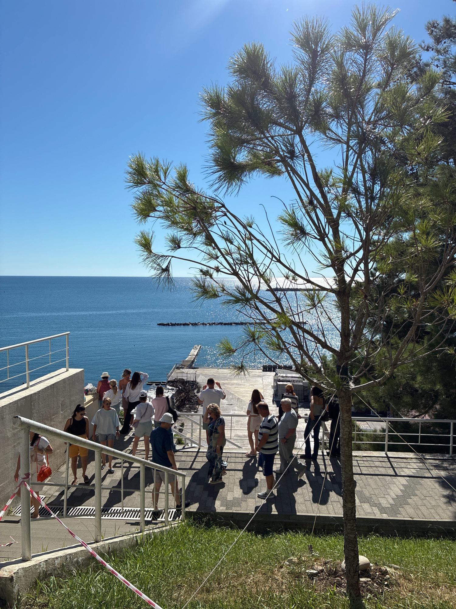 The image depicts a sunny day in Sochi, Russia, overlooking the Black Sea. A group of people are seen walking down a paved staircase leading towards a wooden pier and the water. The sea is calm and blue, with a distant breakwater visible. Lush green trees frame the scene, with one prominent pine tree in the foreground casting a shadow. The people appear to be tourists or locals enjoying a day out, with some dressed in casual summer attire. The overall atmosphere is relaxed and vacation-like.