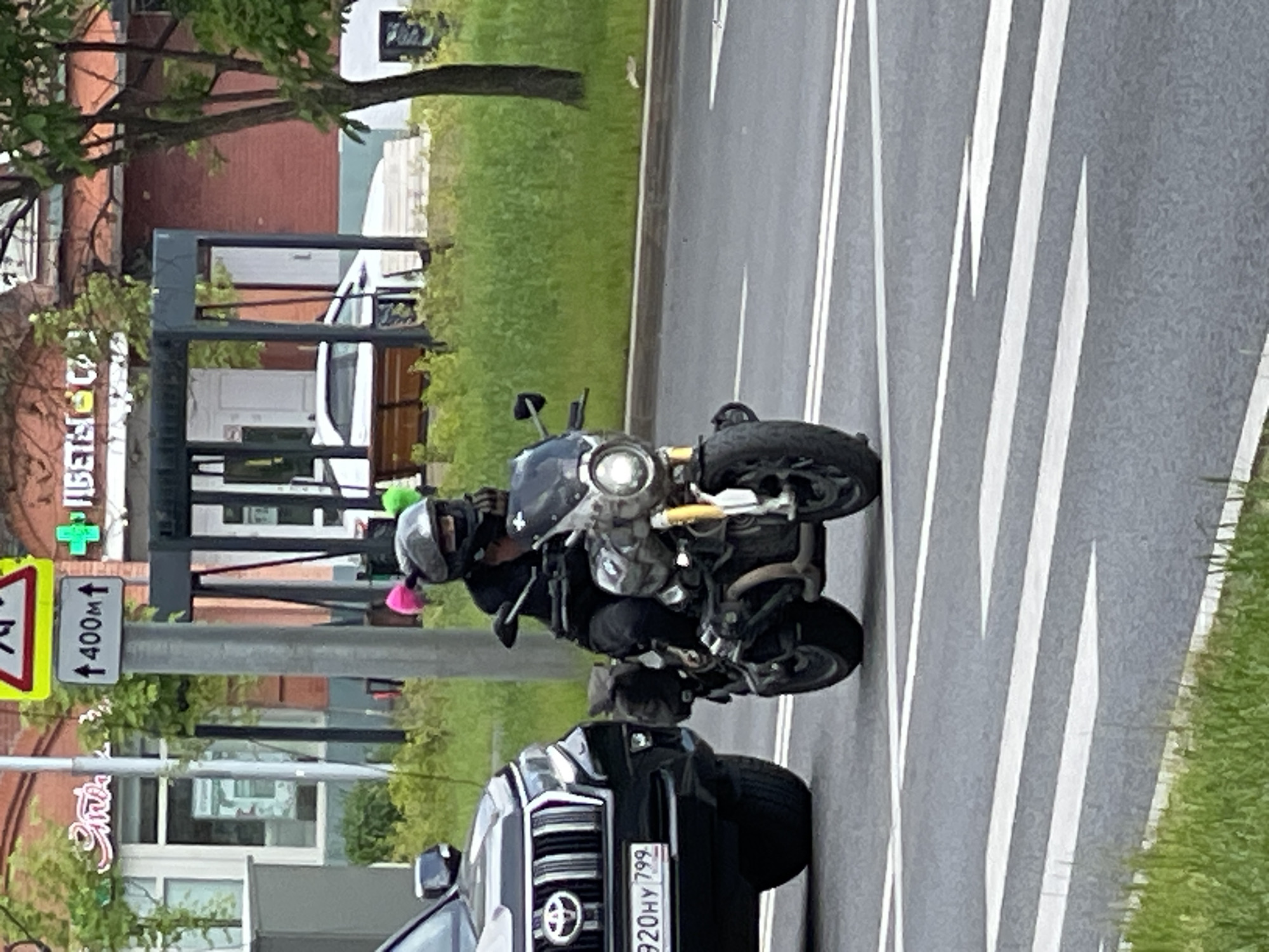 A motorcycle with a rider wearing a helmet and a black jacket is in the foreground, moving from left to right. The motorcycle has a round headlight and a small windshield. The rider has pink fuzzy accessories attached to the helmet. To the left of the motorcycle is the front of a black SUV. The SUV has a license plate that reads "920HY 799". The scene takes place on a paved road with white lane markings. In the background, there is a crosswalk, a bus stop shelter with wooden benches, a brick building with windows, and trees with green foliage. A street sign indicating "400m" with an upward arrow is visible. The environment appears to be an urban setting in Moscow, Russia.