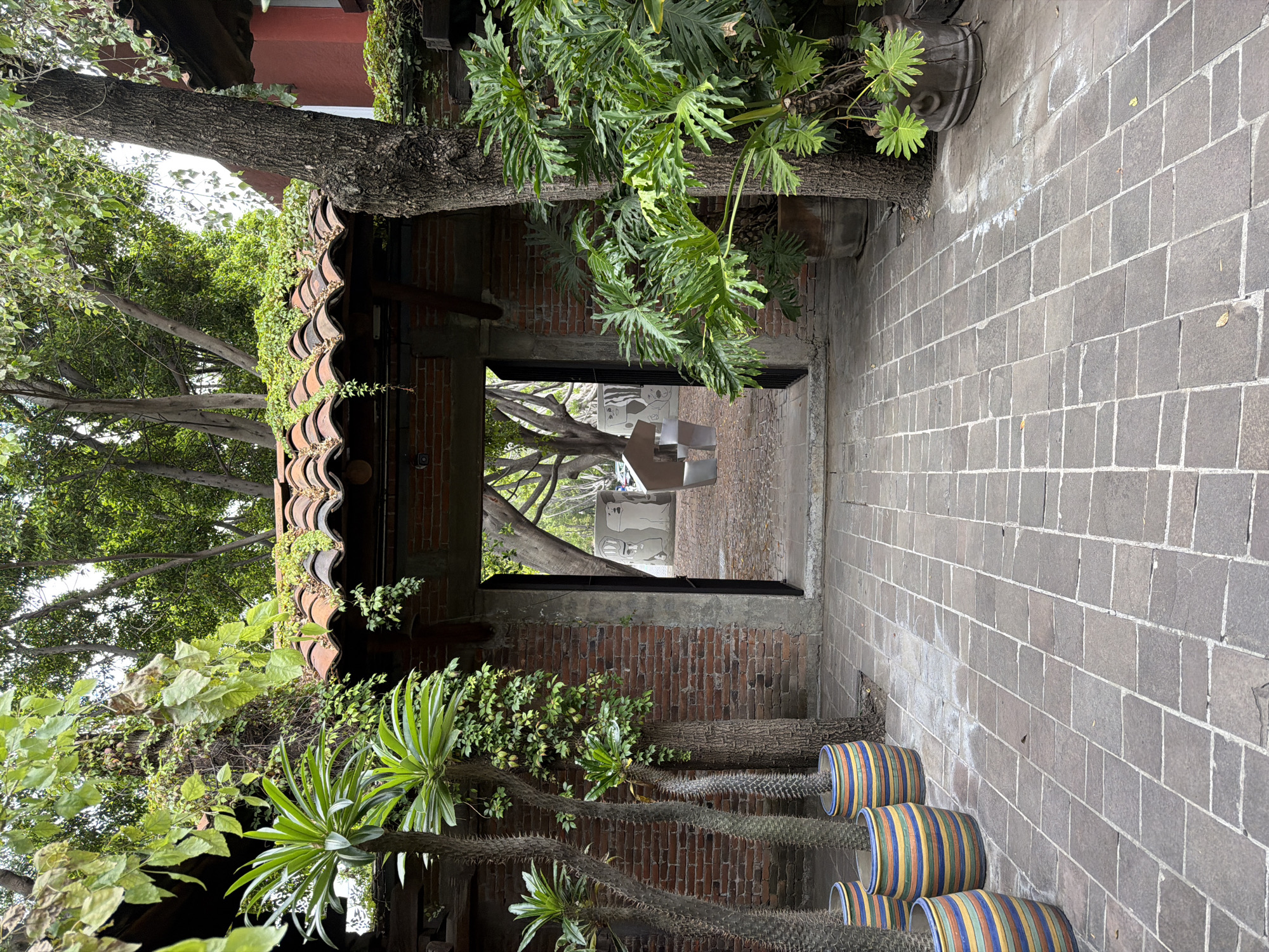 A brick-paved alleyway in Puebla, Mexico, is framed by a brick archway covered in vegetation. Through the archway, a shaded area with a tree and what appears to be art installations is visible. Along the right side of the alley, a row of striped ceramic pots is stacked against the wall, housing spiky plants. The architecture and the plant life suggest a garden or courtyard setting.