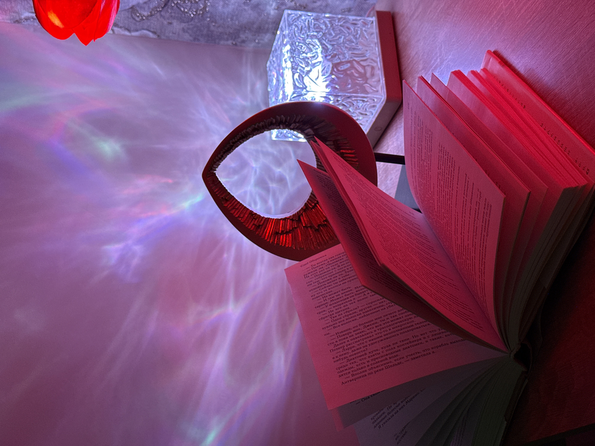 Here is a detailed description of the image:
**Scene Overview:**
The image captures a scene indoors with a book as the focal point, illuminated by multiple light sources creating a spectrum of colors. The overall setting suggests a cozy or artistic environment, possibly in a private residence.
**Subjects and Objects:**
* **Book:** A large book is prominently displayed, partially open. The pages show text in what appears to be a Cyrillic script.
* **Decorative Lighting:** Multiple light sources contribute to the scene's ambiance. A red, tulip-shaped lamp is visible at the top, casting a warm red light. Adjacent to the book is a modern, cubic lamp with a textured surface, emitting a cooler, white light. There are also light patterns on the walls.
* **Abstract Red Decoration:** A red, looped sculptural piece sits near the cubic lamp.
**Environment:**
The background appears to be a soft, possibly padded wall, covered in light and shadows. The light patterns create a colorful, ethereal effect on the wall, suggesting the use of patterned lighting or projections.
**Color and Lighting:**
The color palette is diverse, ranging from deep reds to cooler blues and purples. The lighting is dynamic and contributes significantly to the overall mood of the scene.
**Summary:**
The image captures an interior space where a book is highlighted by a complex interplay of artificial lighting. The setting is artistic and atmospheric.