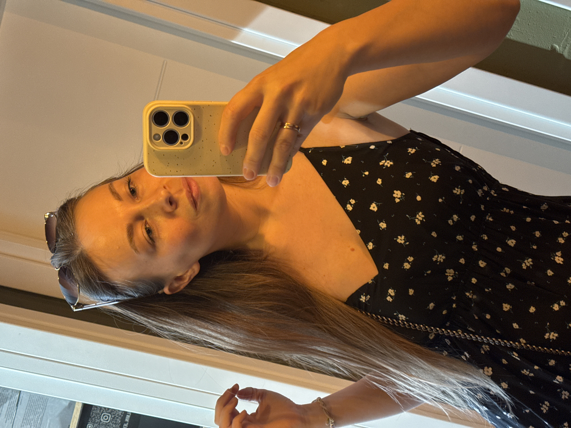A woman is taking a selfie in a mirror. She is wearing a black dress with small white floral patterns and a gold ring on her finger. She is holding a yellow cell phone in her hand. Sunglasses are perched on top of her head. A gold chain is visible on her dress, suggesting it's either part of the dress design or a chain bag strap. The reflection indicates she may be indoors, with a white wall and potentially a window or bright light source nearby. The exact location in Budva, Montenegro, is not discernible from the provided image.
