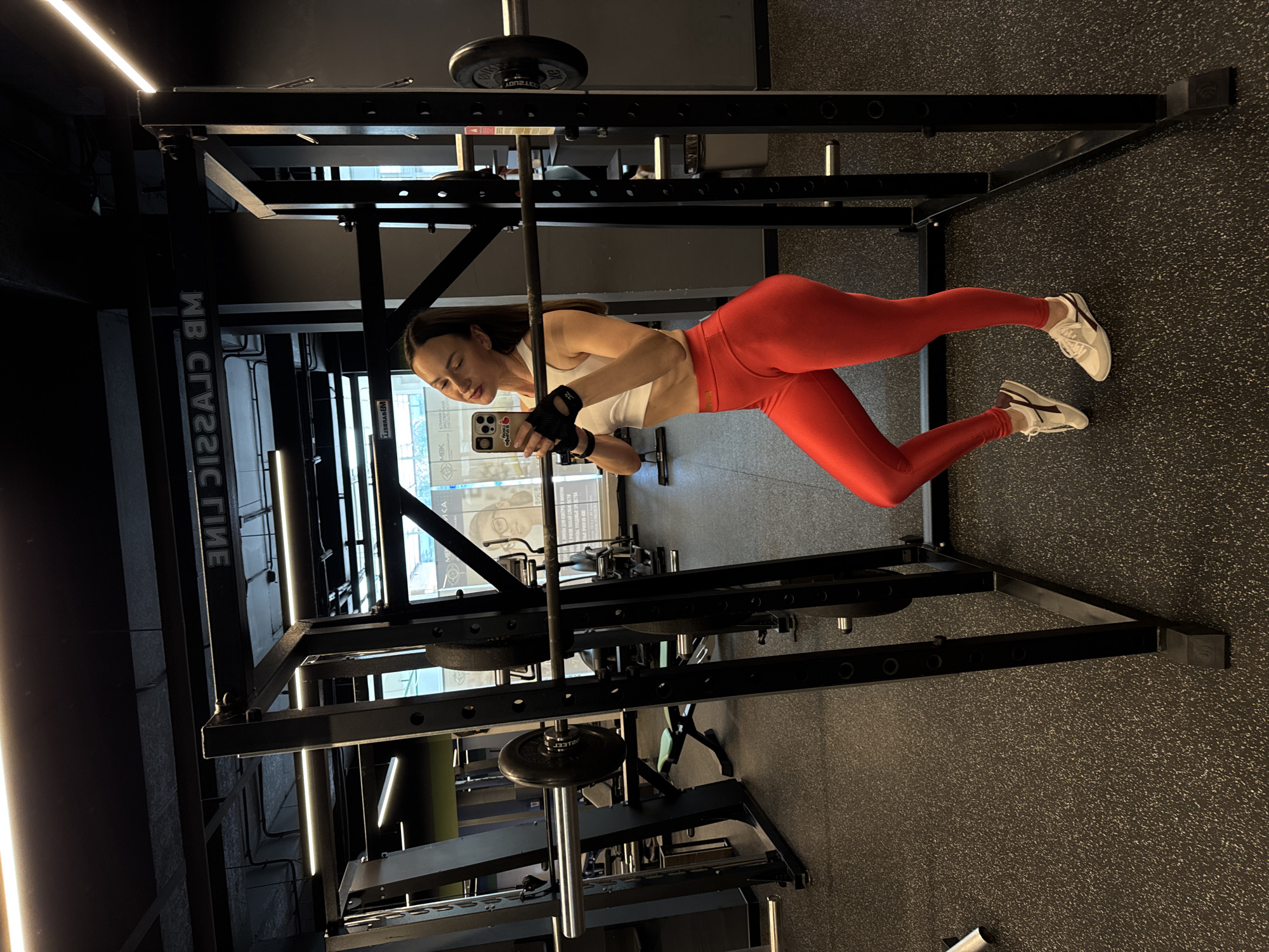 The image captures a fit young woman in a modern gym setting, oriented as if she is floating horizontally due to a 90-degree counter-clockwise rotation of the photograph. When viewed upright, she stands in front of a black power rack, holding a smartphone to take a mirror selfie or video. She has long, dark hair and wears a white sports bra, red high-waisted leggings, white athletic shoes with maroon accents, and black fingerless workout gloves. Her expression is focused and serene.
A barbell, loaded with at least one 20kg weight plate, rests on the safety catches of the power rack. The gym features dark walls, bright linear LED lighting on the ceiling and possibly walls, and a black rubberized floor speckled with white. In the background, through what appears to be a large window or glass partition, reflections show parts of the building exterior or another interior space.
Visible text includes "WB CLASSIC" reversed on a wall or mirror, hinting at the gym's branding. On the back of her smartphone case, a red heart with white text "Я ❤️" (likely part of "Я люблю" meaning "I love" in Russian) is visible, supporting the context of the image being from Sochi, Russia. A small "32" is also seen on her glove. The overall impression is one of fitness, self-documentation, and a well-equipped gym environment.