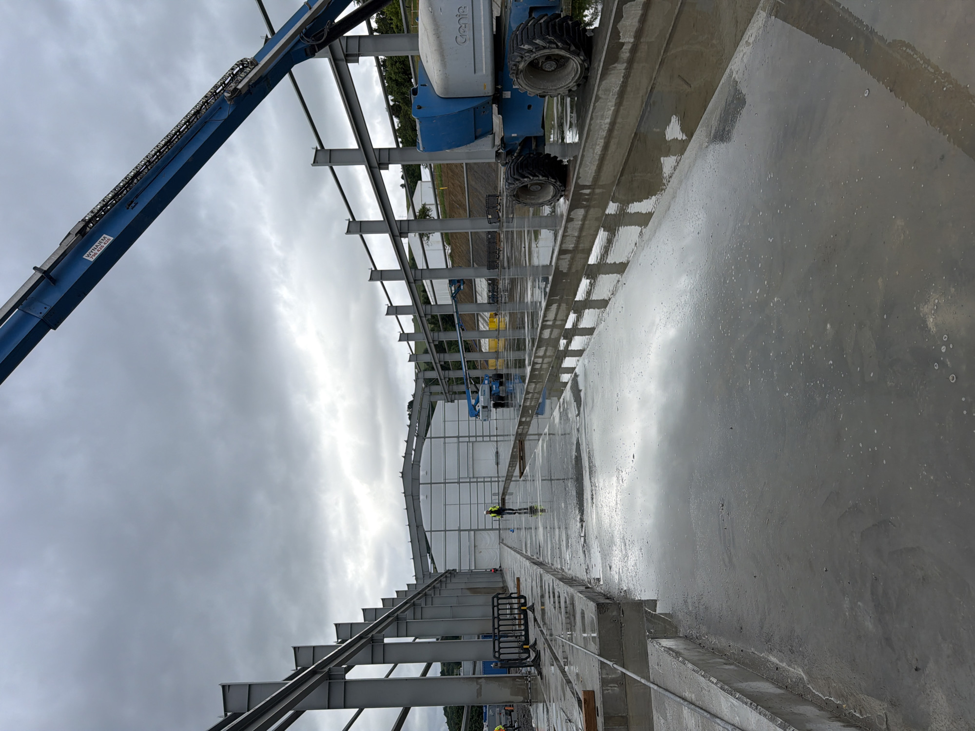 The image depicts a construction site in Stróża, Poland, under an overcast sky. The foreground features a wet concrete surface reflecting the surrounding steel structures of an unfinished building. In the mid-ground, several workers in high-visibility vests are present. One worker, wearing a yellow vest and white hard hat, is walking away from the camera. Another worker, wearing a yellow vest and possibly a dark hard hat, is visible further in the distance near a black safety barrier. A blue aerial work platform, identified as a Genie lift, is parked on the right side of the image, near the large white tent-like structure that forms the rear wall of the building under construction. The overall scene is indicative of ongoing construction activity on a cloudy day.