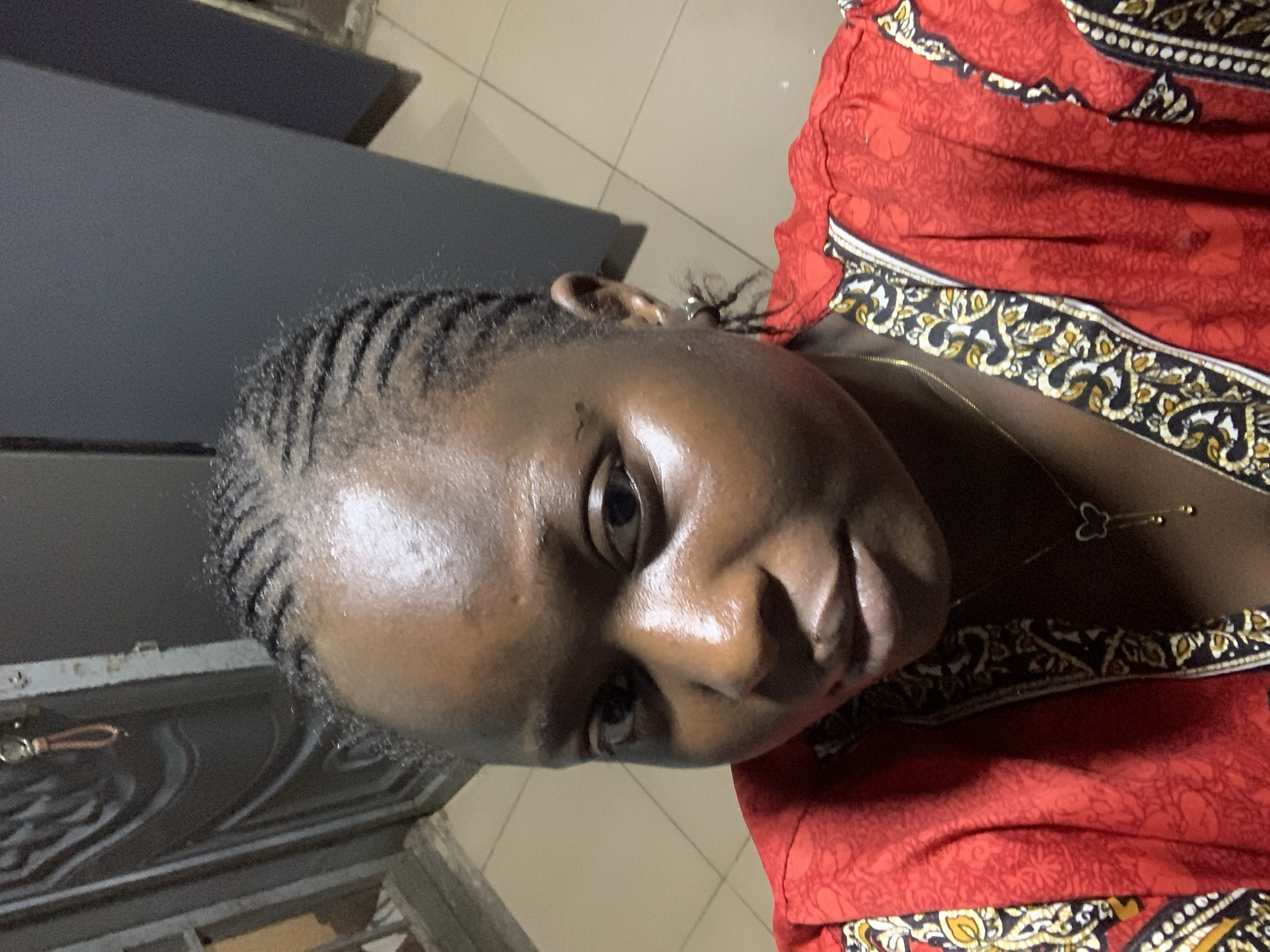 A woman with braided hair and wearing a red and black patterned top is pictured in a close-up selfie. She has a butterfly necklace on. The background appears to be an indoor setting with tiled flooring and a dark gray wall.