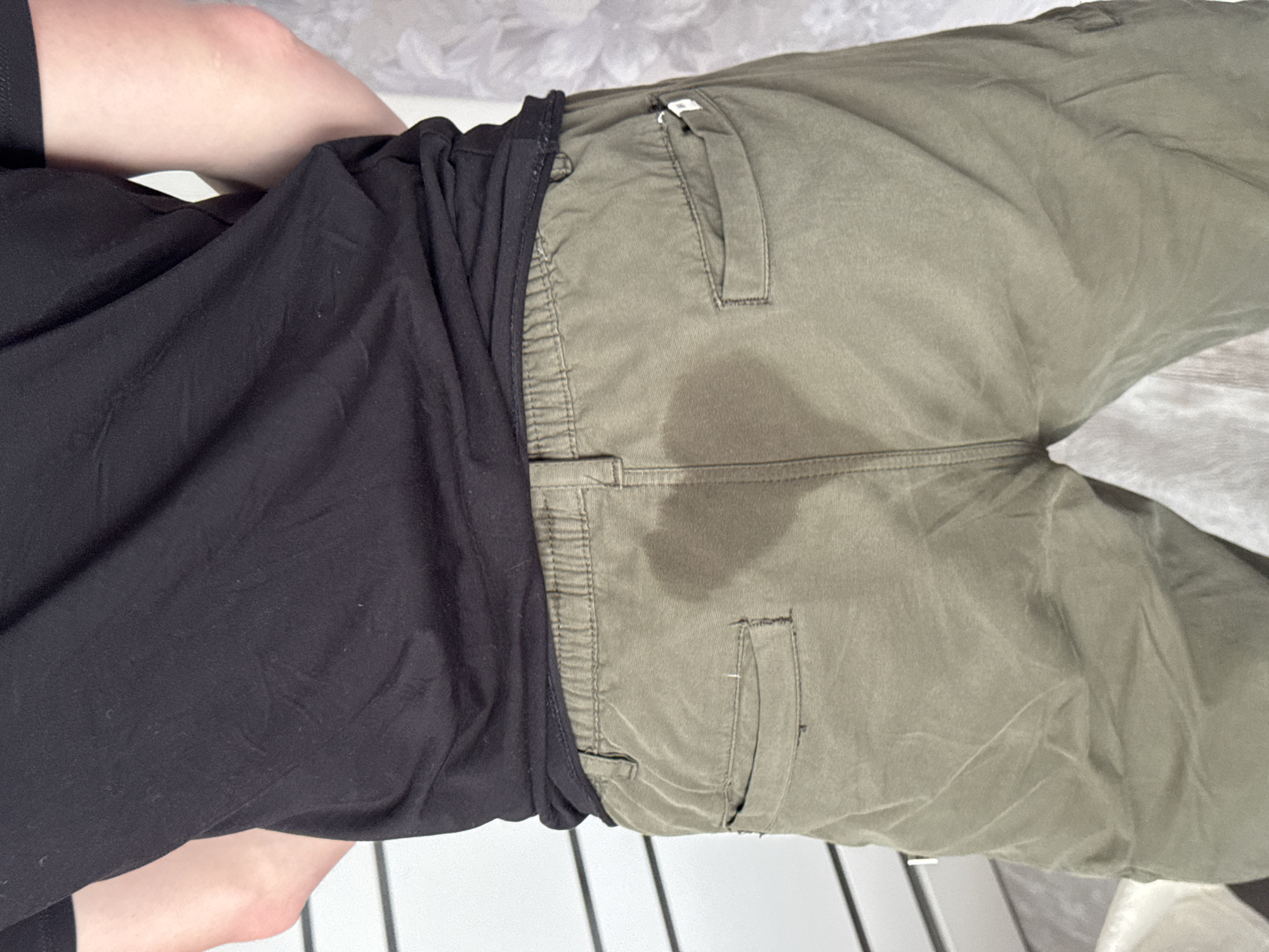 The image shows a person lying down, wearing black shorts and olive green trousers. The trousers have a darker wet patch in the crotch area. The setting appears to be indoors, with white flooring and a patterned surface behind the person's head. There is no information in the image to indicate this image was taken in Krasnodar, Russia.