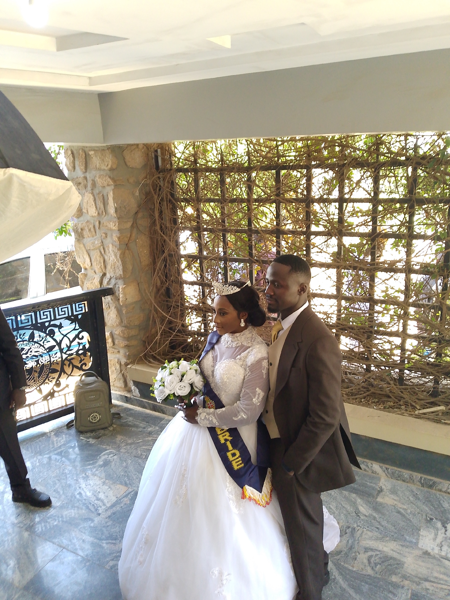 The image depicts a bride and groom posing for a photograph in an outdoor setting, possibly a patio or covered area of a building. The bride is wearing a white wedding gown with lace details, a tiara, and a sash that reads "BRIDE." She holds a bouquet of white flowers. The groom stands next to her, wearing a brown suit and a light-colored tie. Behind them is a wall with a lattice covered in vines. To the left, part of a photography light with a softbox is visible. A person in a suit is partially visible on the left edge of the image. There is a backpack on the floor behind the bride. The floor is covered in marble. The location appears to be in the city of Unknown, Unknown.
