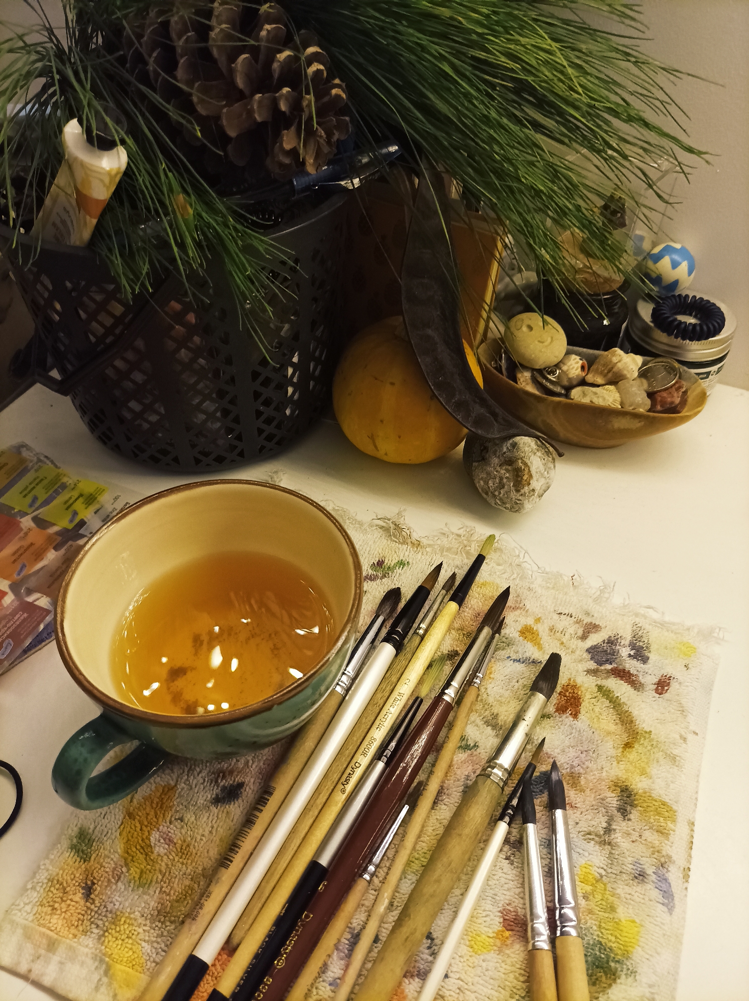This image depicts an indoor tabletop or desk scene located in Batumi, Georgia. In the immediate foreground, a teal ceramic mug with a light brown rim holds an amber-colored liquid containing small dark specks. Adjacent to the mug, approximately 15-20 assorted paintbrushes, varying in size and handle material, are laid horizontally across a small, textured, off-white towel. The towel exhibits multiple paint stains in colors such as blue, yellow, green, and brown.
To the left of the mug, a portion of a blister pack containing small paint tubes or watercolor pans is visible. In the midground, a small, spherical orange object, resembling a gourd or pumpkin, is situated next to a dark, curved, elongated seed pod. A small wooden bowl holds various items including seashells, small stones (one marked with a smiley face), crystalline fragments, and small metallic objects. Behind the bowl, a clear glass container is present. To the right, a small blue and white patterned ball and a dark blue coiled hair tie resting on a silver-lidded jar are visible.
The background on the left features a dark, perforated container holding a cluster of dark green pine needles and multiple large pinecones. A partially used tube of cream-colored paint with orange details is nestled within the pine needles. The background wall is plain and light-colored.