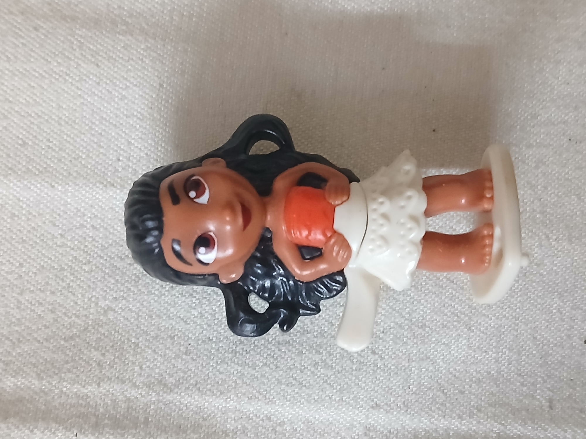 A Moana doll is lying on a white, textured surface. The doll is made of plastic and has dark hair, brown skin, and is wearing a white skirt and orange top. The doll is lying on its back. The location is likely Bengaluru, India.