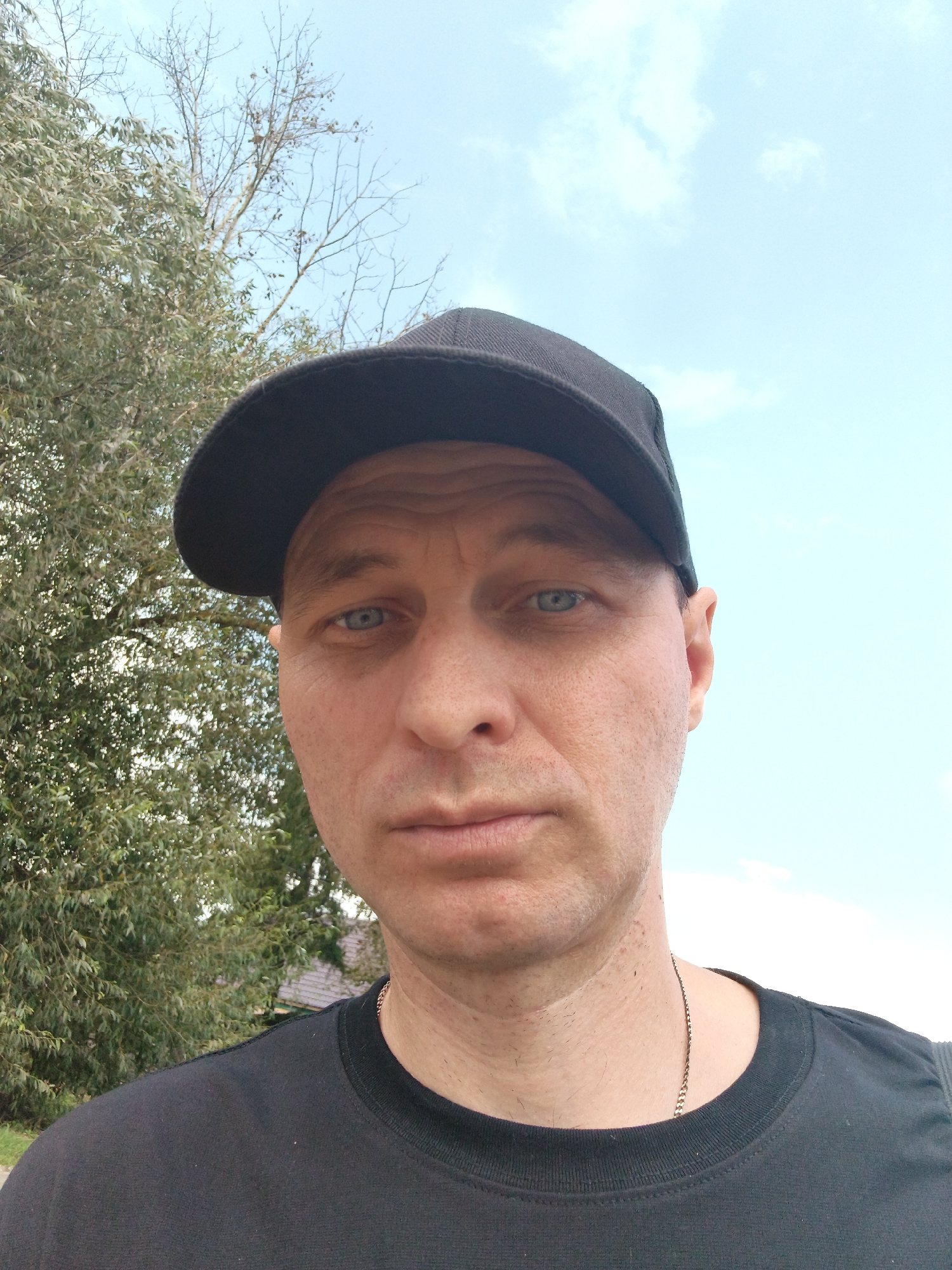 A man with fair skin, short brown hair, and blue eyes is captured in a close-up, selfie-style photograph. He is wearing a black baseball cap and a black t-shirt with a thin silver chain around his neck. His expression is neutral, and he is looking directly at the camera. The background consists of green foliage from a tree on the left side and a bright blue sky with scattered clouds on the right. The image appears to have been taken outdoors, possibly in a park or residential area, given the presence of the tree and what seems to be a roof in the lower left portion of the background. The man's facial features, including wrinkles around his eyes and across his forehead, suggest he is middle-aged. There are no other people or significant objects in the frame. The lighting is natural, indicating it's daytime. The image does not contain any visual cues that specifically place it in Mokshan, Russia.