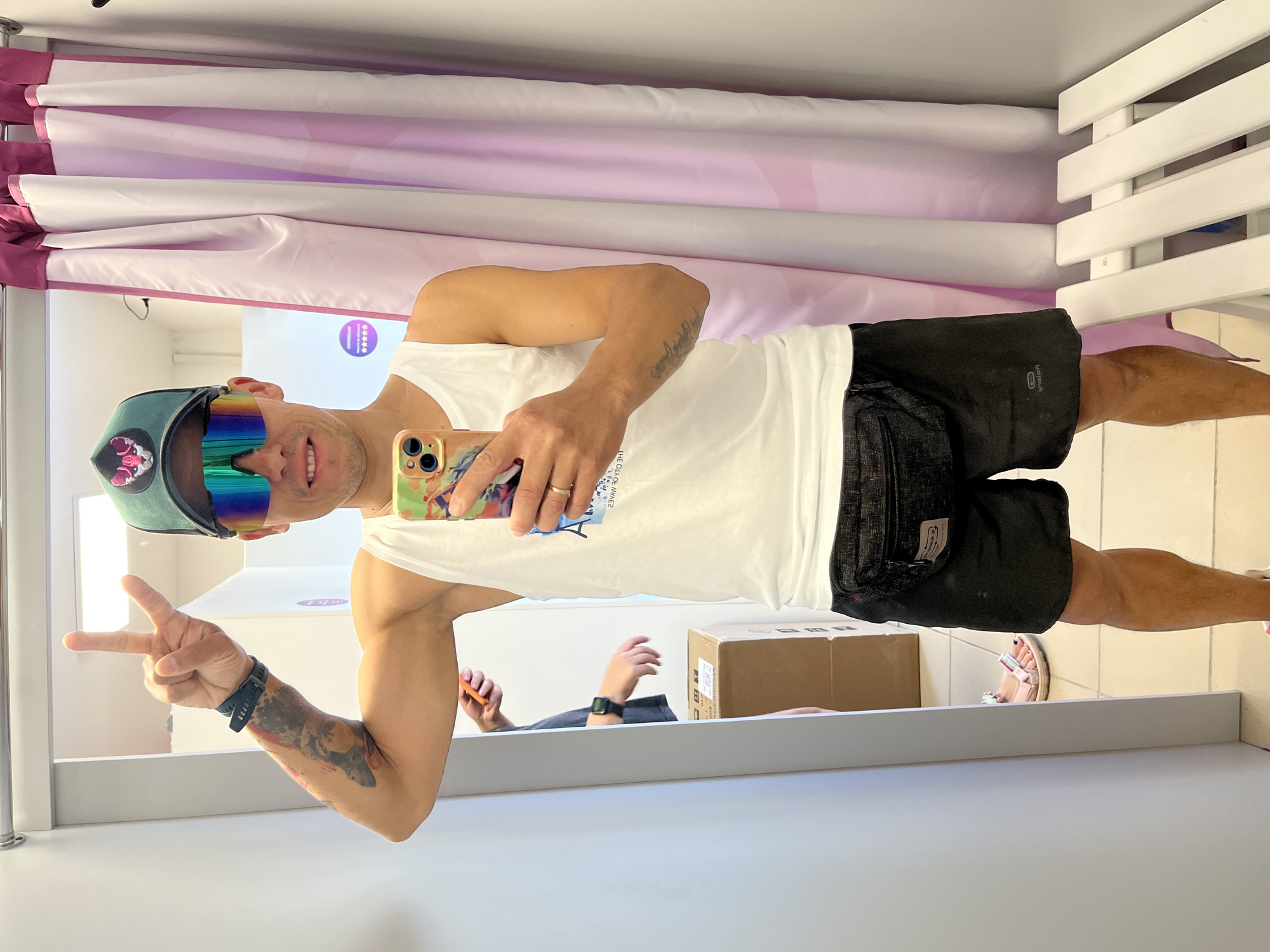 A man wearing a white tank top, black shorts, a green baseball cap, and iridescent sunglasses poses for a selfie in what appears to be a fitting room. He is holding a smartphone in his right hand and making a peace sign with his left hand. A fanny pack is worn around his waist. In the background, a person wearing a grey t-shirt is visible, holding a mobile phone, and two cardboard boxes are stacked on the floor. A pink curtain hangs to the right of the frame. A white bench is positioned in the lower right corner. The scene is set in Chaikovsky, Russia.