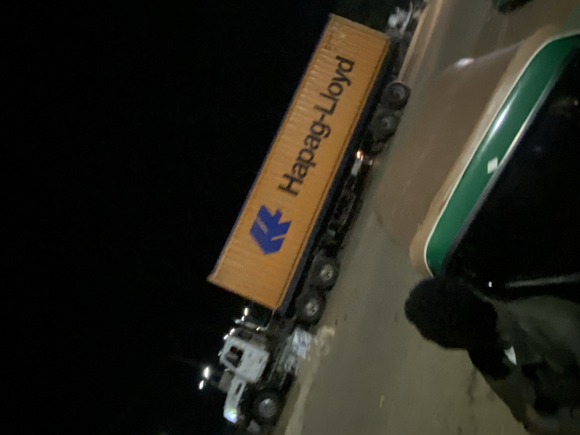 In the city of Bukuru, Nigeria, at night, a white semi-truck carrying an orange Hapag-Lloyd shipping container is parked on a paved surface. The truck is positioned at an angle, with its front facing left and the container extending to the right. The shipping container has a blue logo and the text "Hapag-Lloyd" printed on its side. In the foreground, a dark-colored vehicle, possibly a bus or van, is partially visible, with a person's silhouette in the driver's seat. The surrounding environment is dark, with artificial lights from the truck and streetlights casting illumination on the scene.