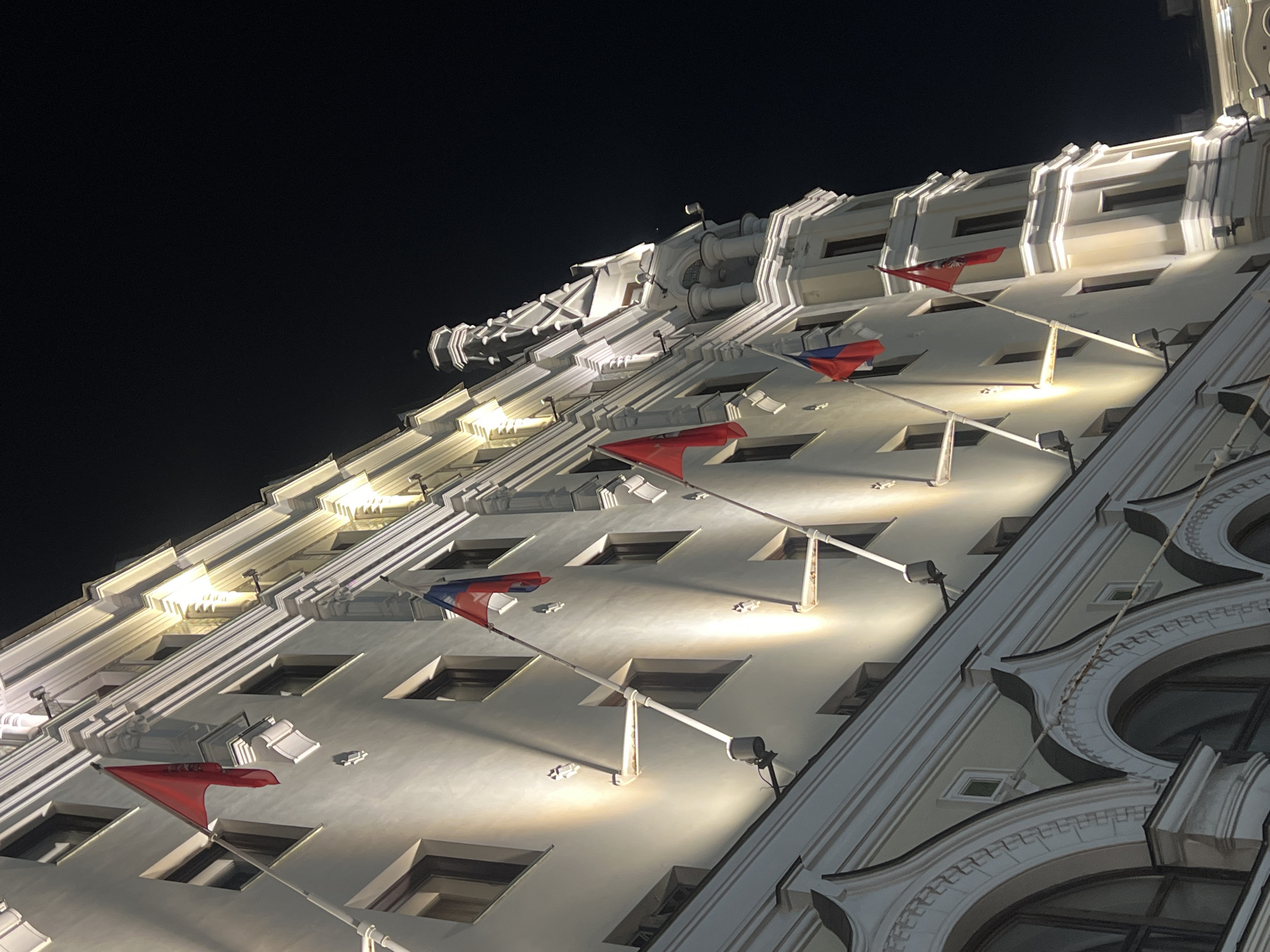 The image presents a detailed view of the illuminated facade of the Moscow City Hall (Мэрия Москвы), located on Tverskaya Street in Moscow, Russia, at night. The grand, multi-story building features a light-colored, possibly cream or light grey, facade with classical architectural elements, including prominent white cornices, decorative moldings, and regularly spaced rectangular windows with dark frames. The visible base of the building is characterized by large, arched windows with intricate white decorative surrounds.
The facade is dramatically lit from below by multiple warm white spotlights, casting strong upward shadows and accentuating the building's intricate details and textures. Several flags are displayed on flagpoles extending diagonally outwards from the building's upper stories. Specifically, from left to right, the flags include a plain red flag, followed by a flag of the Russian Federation (horizontal white, blue, and red stripes). Further to the right are additional flags: a red flag bearing the golden emblem of Moscow (Saint George slaying the dragon), another plain red flag, another Russian Federation flag, and a final red flag with the Moscow emblem.
On the far right, the building's structure culminates in an ornate, multi-tiered turret or tower featuring columns, arches, and intricate stonework, all similarly illuminated. The sky above is entirely dark, indicating the nighttime setting. There are no visible people or vehicles within the frame, focusing solely on the architecture and its nocturnal illumination.