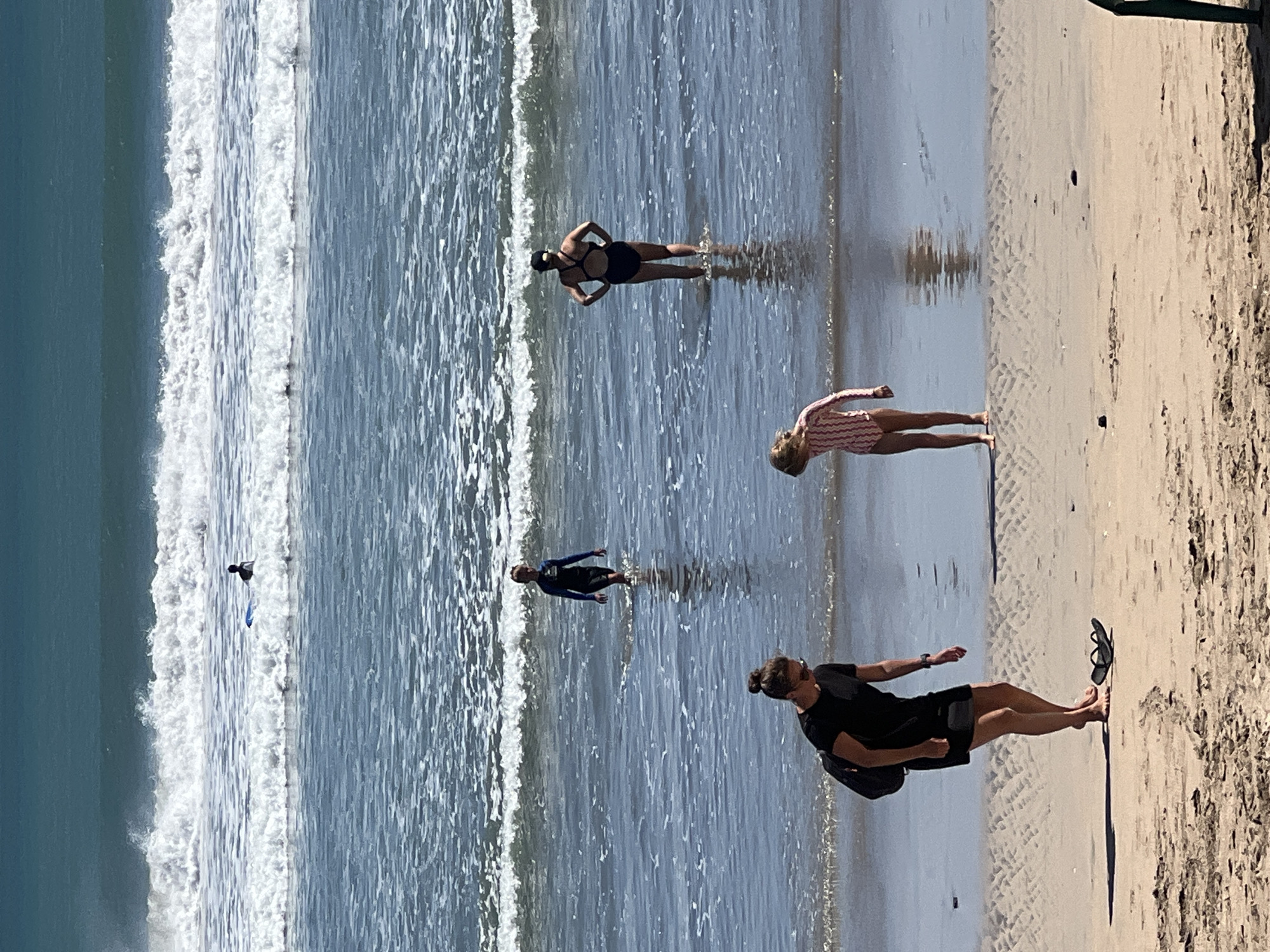 In Kuta, Indonesia, a beach scene unfolds with several individuals enjoying the ocean. In the foreground, two women and a young girl are on the sand. One woman, wearing a black dress and carrying a backpack, stands barefoot near a pair of black flip-flops. A young girl in a striped swimsuit stands a few feet away, looking down. Further out in the shallow water, two other women are present. One, in a black swimsuit, stands with her hands on her hips, facing away from the camera. Another person, also in a dark swimsuit, walks through the water towards the shore. In the background, a lone surfer on a blue surfboard sits in the waves, with more breaking waves further out and the turquoise ocean stretching to the horizon under a bright sky.