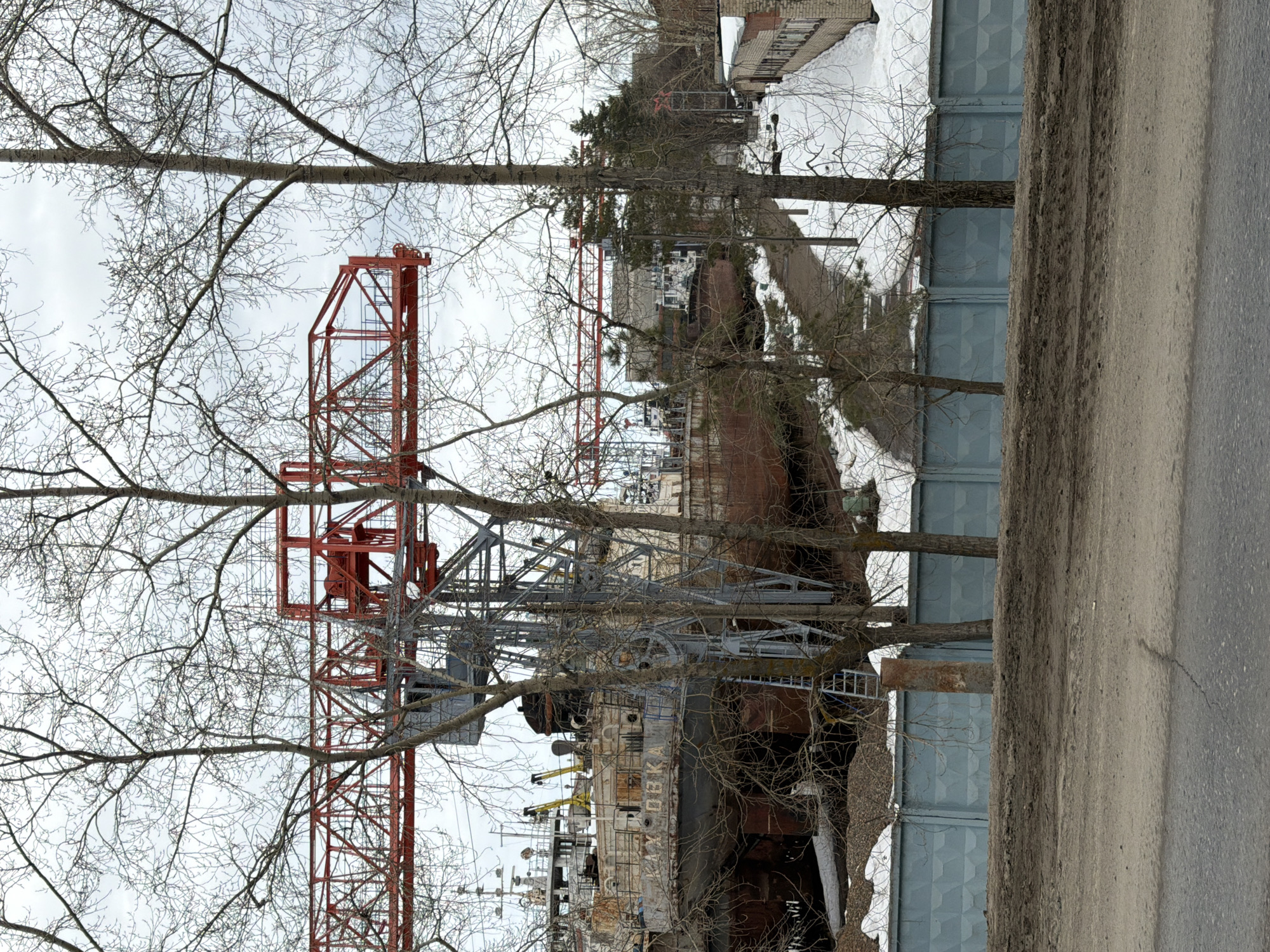 The image depicts a construction crane towering over what appears to be a shipyard or ship repair facility. The crane is red and gray and is partially obscured by bare tree branches. The facility contains structures including ship hulls. The location is likely in Chaikovsky, Russia, based on the metadata. The scene is overcast with signs of snow, and a street is visible on the right.