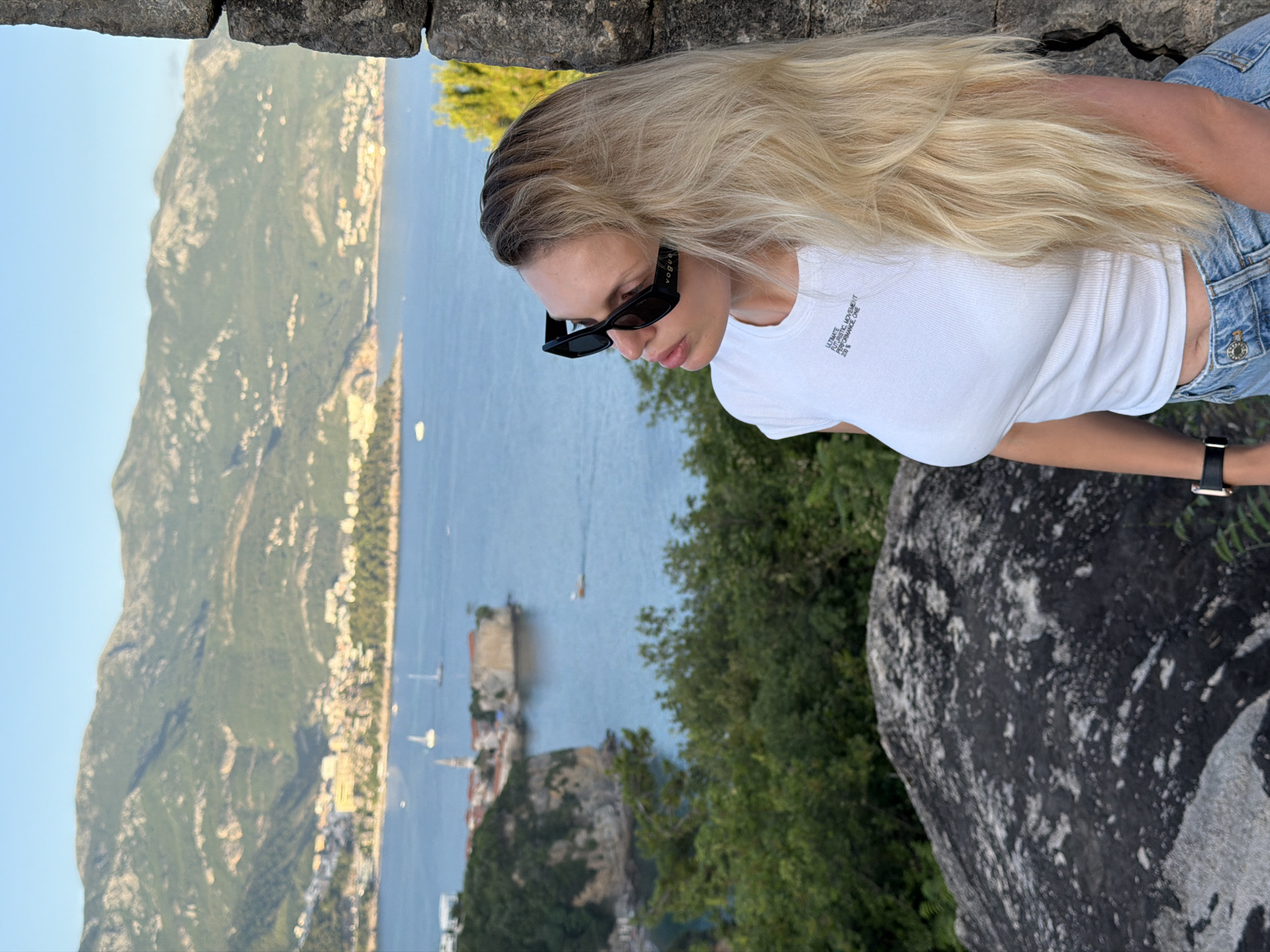 A young woman with long blonde hair and black sunglasses stands leaning against a stone wall, looking out at a scenic coastal view. She is wearing a white t-shirt with text and blue denim shorts. The background features the blue sea, a city nestled along the coastline with buildings and a church steeple, lush green hills, and mountains in the distance. Boats are visible on the water, including sailboats and a small motorboat. The scene appears to be set in the city of Budva, Montenegro, likely from a vantage point overlooking the Old Town and the Adriatic Sea. The lighting suggests it is late afternoon or early evening, with a clear sky.