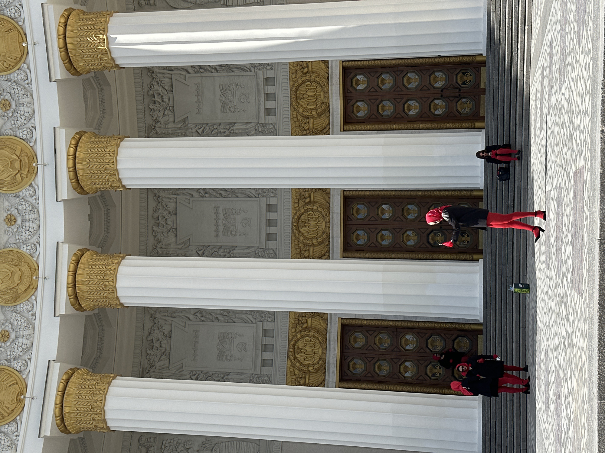 The image depicts an architectural facade with prominent columns and ornate detailing, potentially located in the city of Unknown, Unknown. The columns are white with gold accents at the top, and they stand in front of a wall with decorative panels and doors with intricate patterns.
Several individuals are visible on the steps in front of the building. They are wearing black clothing with striking red tights and hats. One person is standing and appears to be in motion, while others are grouped together near the bottom of the steps. The setting appears to be outdoors during daylight, based on the lighting and shadows.