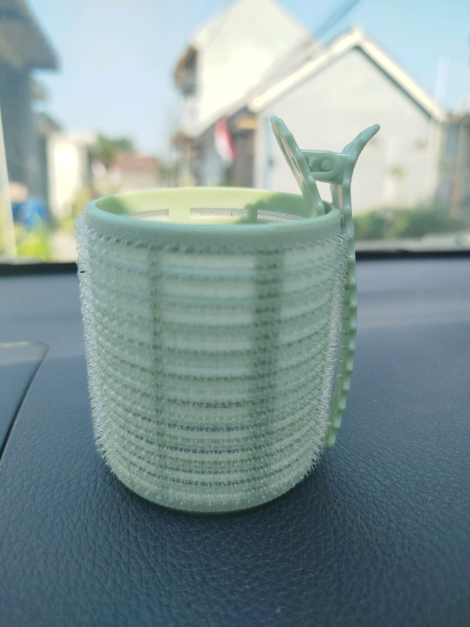 A close-up, eye-level shot shows a light green hair roller with a clip attached to it, placed on a dark gray textured surface, likely the dashboard of a car. The roller has a ridged texture and a fuzzy outer edge. In the background, through what appears to be a car windshield, a blurred outdoor scene is visible. This includes a white building with a gabled roof, a few other structures, and some greenery. The sky above is a clear, light blue. The setting is consistent with a car parked or driving in the area of Cagakagung, Indonesia, under daylight conditions.
