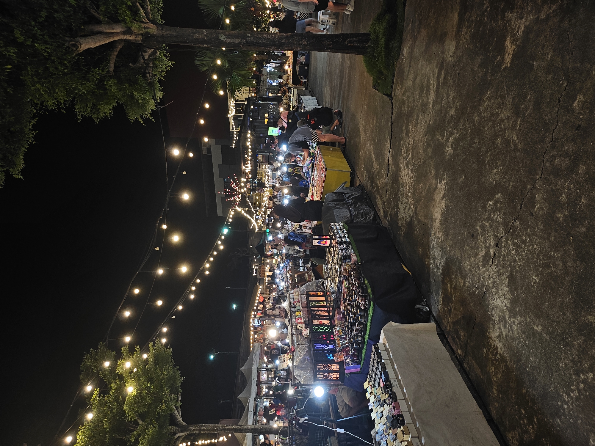 The image depicts a vibrant night market in Phuket City Municipality, Thailand, illuminated by strings of warm, glowing bulbs that crisscross the scene, creating a festive atmosphere. Numerous stalls, some covered by white awnings, display a variety of goods, including what appear to be decorative items, small boxes of merchandise, and possibly soaps or cosmetics. People are scattered throughout the market, some browsing the stalls, others engaged in conversations, and a few are working behind the counters. The ground is paved with a rough, textured concrete surface. Tall trees with lush green foliage are visible, adding to the outdoor setting. In the background, buildings with lights suggest a more urban environment beyond the market. The overall scene captures the lively energy of a popular local marketplace at night.