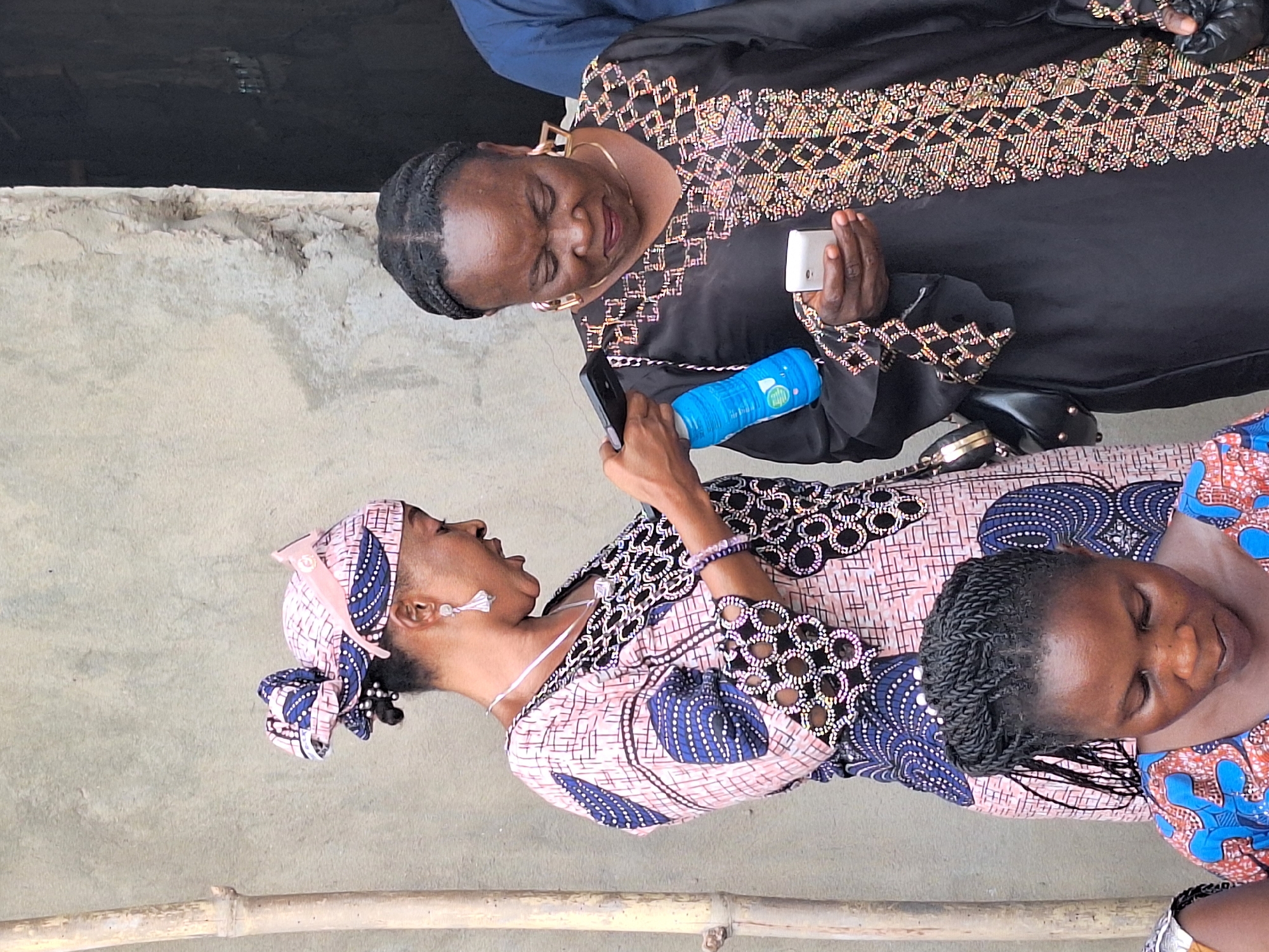 In an outdoor setting, likely in Sabon Tasha, Nigeria, three women are present. One woman, wearing a black gown with gold and silver embroidery, holds a white smartphone in her right hand and a blue cylindrical object in her left. She is facing left and looking down at a woman with a headscarf. The woman in the headscarf, wearing a dress with pink, blue, and black patterns, is holding a black smartphone and looking up at the woman in the black dress. To her right, a third woman with braided hair is partially visible, wearing a blue and orange patterned dress. The background consists of a gray concrete surface and a horizontal bamboo structure.