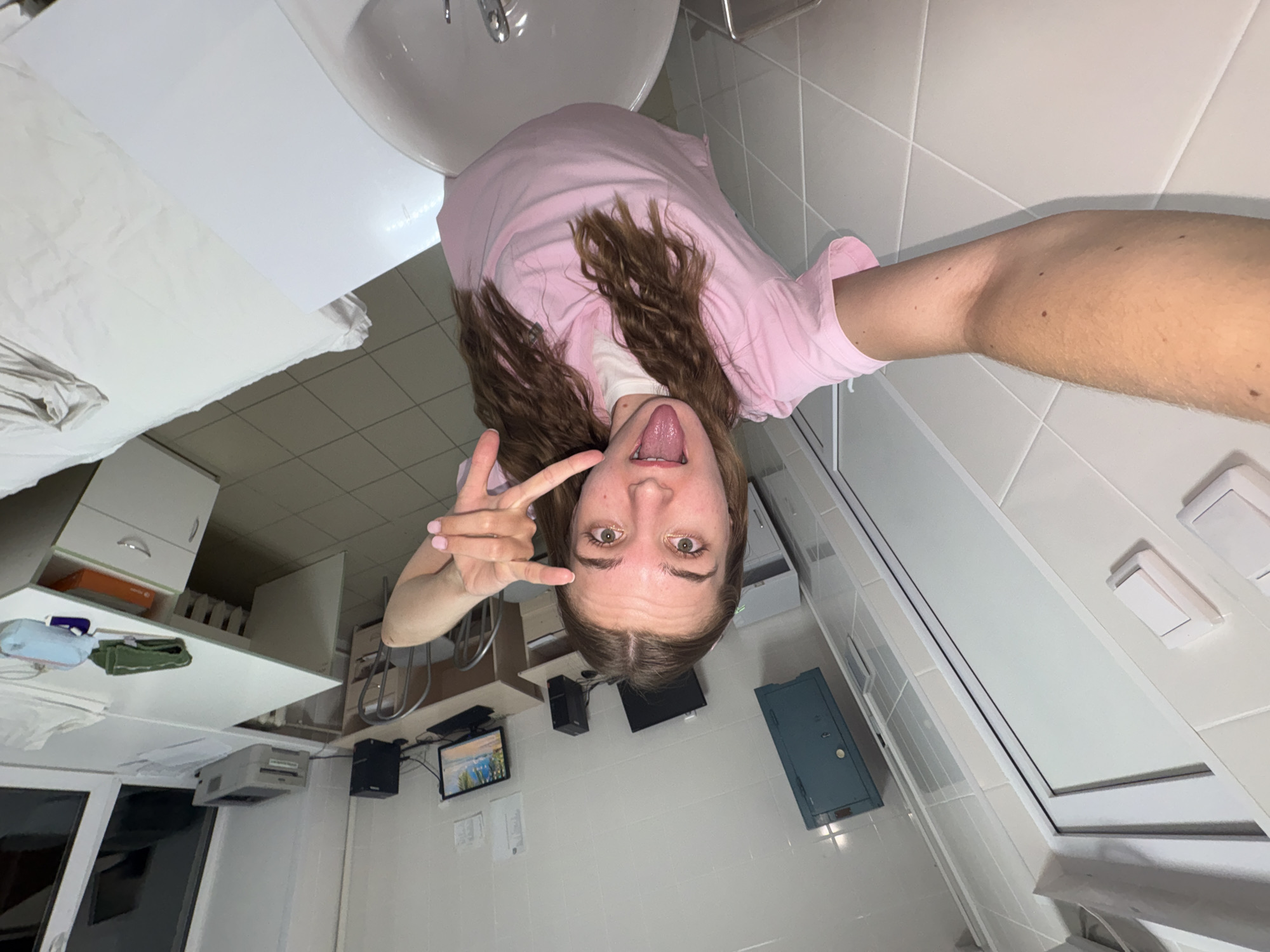 A young woman with long brown hair is taking a selfie in what appears to be a medical or laboratory setting. She is wearing a pink scrub top over a white t-shirt and is sticking out her tongue while making a peace sign with her right hand. Her left arm is visible, extended to hold the camera. The background includes white tiled walls, a light blue metal cabinet mounted on the wall, a desk with computers and a printer, a sink, and a bed covered with white linens. The lighting suggests an indoor environment. The scene is captured from a high angle, looking down at the woman. There are no specific identifiers in the image that would place it in the city of Perm, Russia.