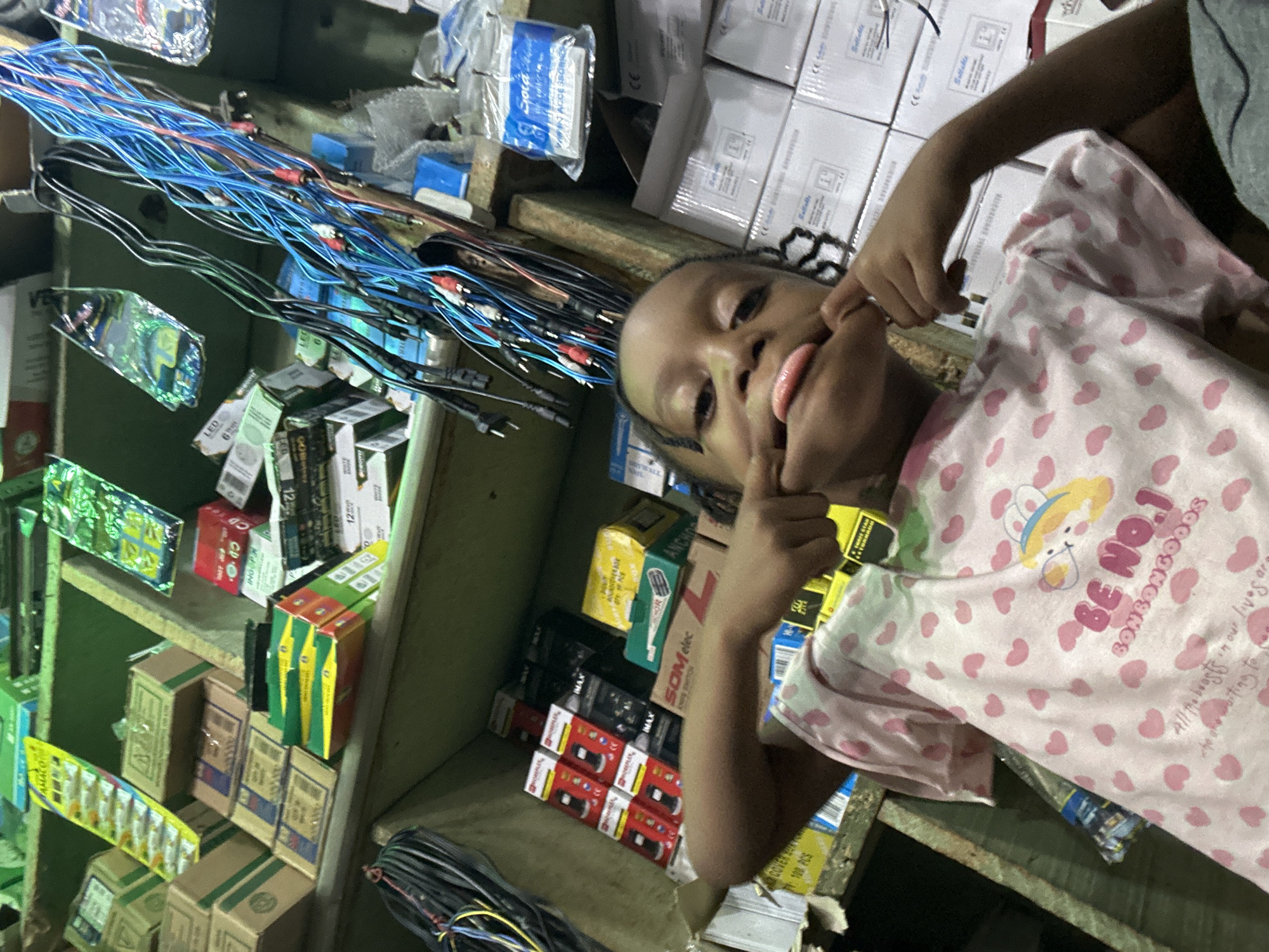 The image depicts a young individual, possibly a child, situated in what appears to be an electronics or hardware store in Narayi, Nigeria. The individual, with dark skin and braided hair, is wearing a light pink t-shirt adorned with small pink hearts and a graphic featuring a cartoon character with text reading "BE NO.1 BONBONGOODS." The child is positioned in the lower center of the frame, looking directly at the camera, and making a playful face by pulling their cheeks outward with both hands and sticking out their tongue.
The background consists of stacked wooden shelves filled with various merchandise. On the upper left shelf, numerous brown and green cardboard boxes are visible, alongside a yellow display card for small items marked "AMACO" and other packaged goods in clear plastic. Below this, on the lower left shelf, are more stacked boxes, including red and white branded boxes with "HOMEPLEX" and "IMAX" labels, and yellow and green boxes featuring "SOM elec" and "ANCHOR" branding. Coiled black and blue electrical cables are visible in the lower left foreground.
To the right, a large bundle of cables, predominantly blue and black, with red and white RCA connectors and other jacks, hangs from the shelf. Below this, several transparent plastic bags, one labeled "Sjora" and "WIRING ACCESSORIES," are visible. Further right, stacks of white cardboard boxes with "Solladie" and "CE" markings suggest electronic components or fixtures. The lighting is artificial, illuminating the goods on the shelves.