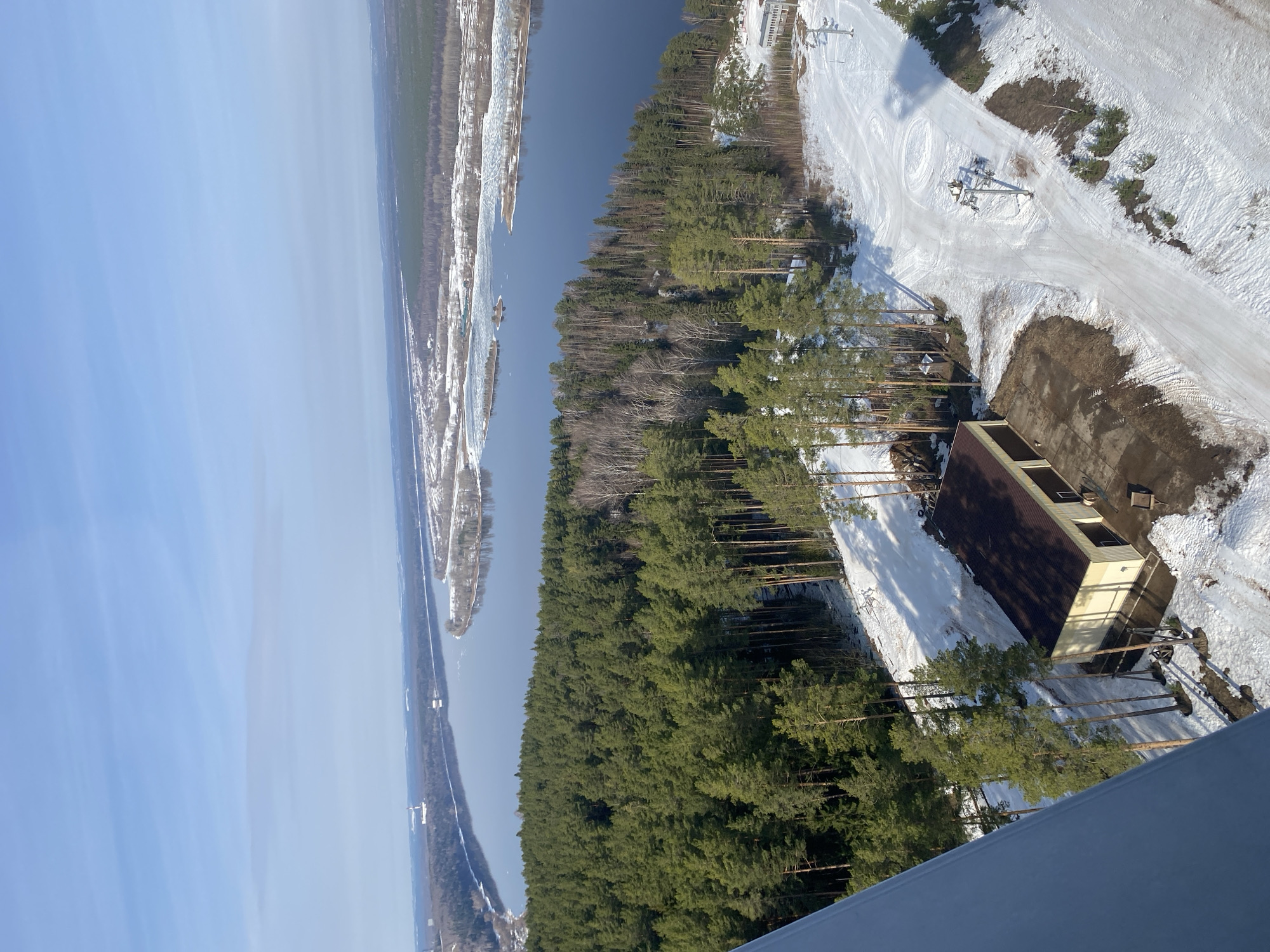 Aerial view of Прикамский, Russia. River to the left with ice. Forest with snow. A ski slope and chairlift. A low, single-story structure is situated at the edge of the forest near the slope.
