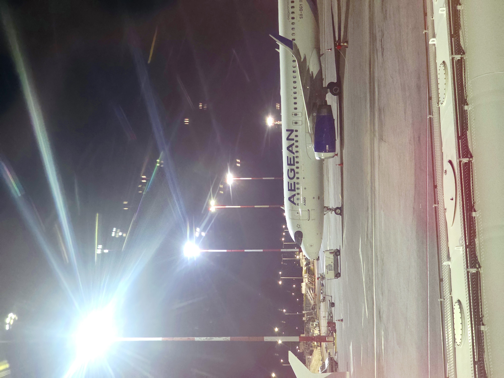 The image depicts an Aegean Airlines airplane parked on the tarmac at night, likely at an airport in Mouzouras, Greece, given the context. The plane, identified as an A320 with registration SX-DGY, is white with the airline's blue logo on the tail and engine cowling. Several bright airport floodlights illuminate the scene, causing lens flare and streaks of light across the dark sky. In the background, other airport infrastructure, including utility vehicles and support equipment, are visible. The foreground shows a section of the airport pavement with markings and a few safety cones. A person in a red uniform can be seen near the rear of the aircraft. The overall atmosphere is one of a nighttime operation at an airport.