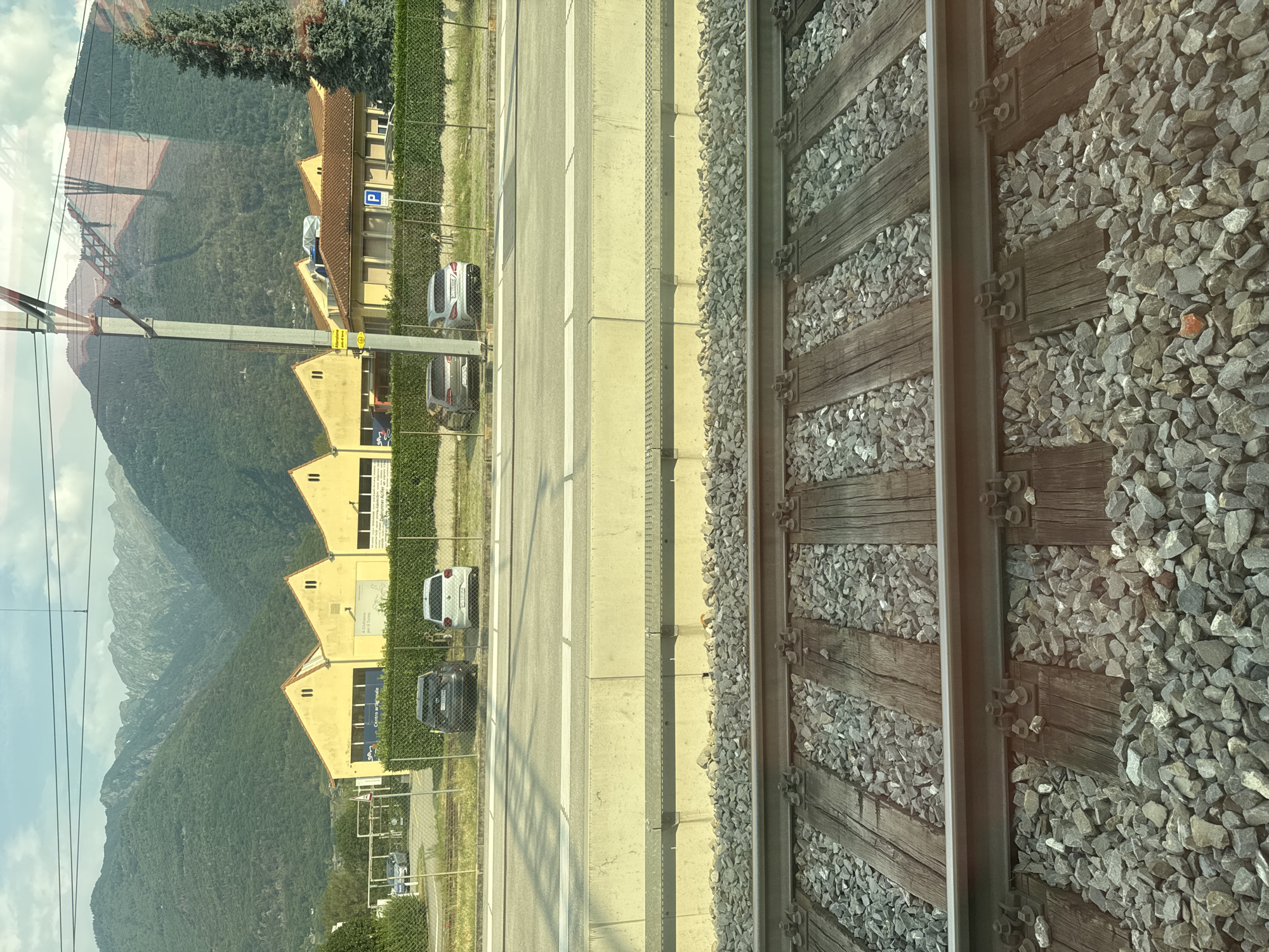 The image depicts a train track in Bellinzona, Switzerland, with a backdrop of mountainous terrain and industrial buildings. The foreground is dominated by the detailed texture of the train tracks and gravel. In the middle ground, a concrete platform separates the tracks from a paved area with parked cars and a fenced-off grassy section. Beyond this, a row of yellow industrial buildings with distinctive gabled roofs is visible, set against the lush green slopes of the mountains. A tall evergreen tree stands to the right of the buildings. The sky above is partly cloudy, with sunlight casting shadows and reflections on the scene, including on the adjacent industrial buildings. There are no people visible in the frame.