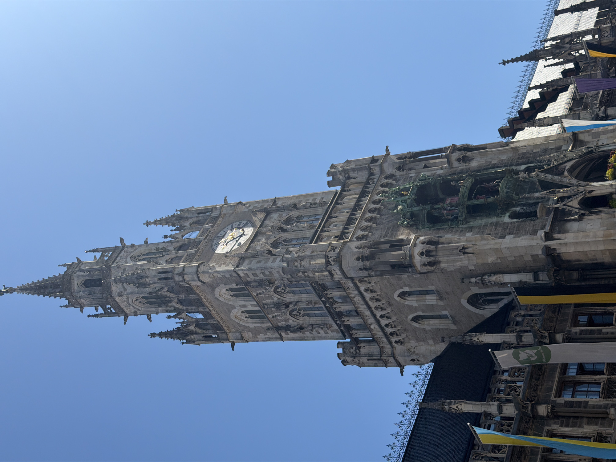 The image shows a low-angle, close-up view of the Neues Rathaus (New Town Hall) in Munich, Germany. The towering Gothic Revival building, constructed primarily of stone, dominates the frame, its intricate spires and facade reaching towards a clear, bright blue sky. A prominent clock face with Roman numerals is visible on the tower, showing approximately 11:40. Below the clock, a large, ornate Glockenspiel featuring animated figures is present. Gargoyles and decorative carvings adorn the building's exterior. At the base of the building, several large, vertical banners are displayed; one is blue and yellow, another is white with a green emblem and text, and a third is black and yellow. The surrounding architecture, including a dark, tiled roof and more pointed spires, is also visible. The scene is devoid of any people or discernible activity.