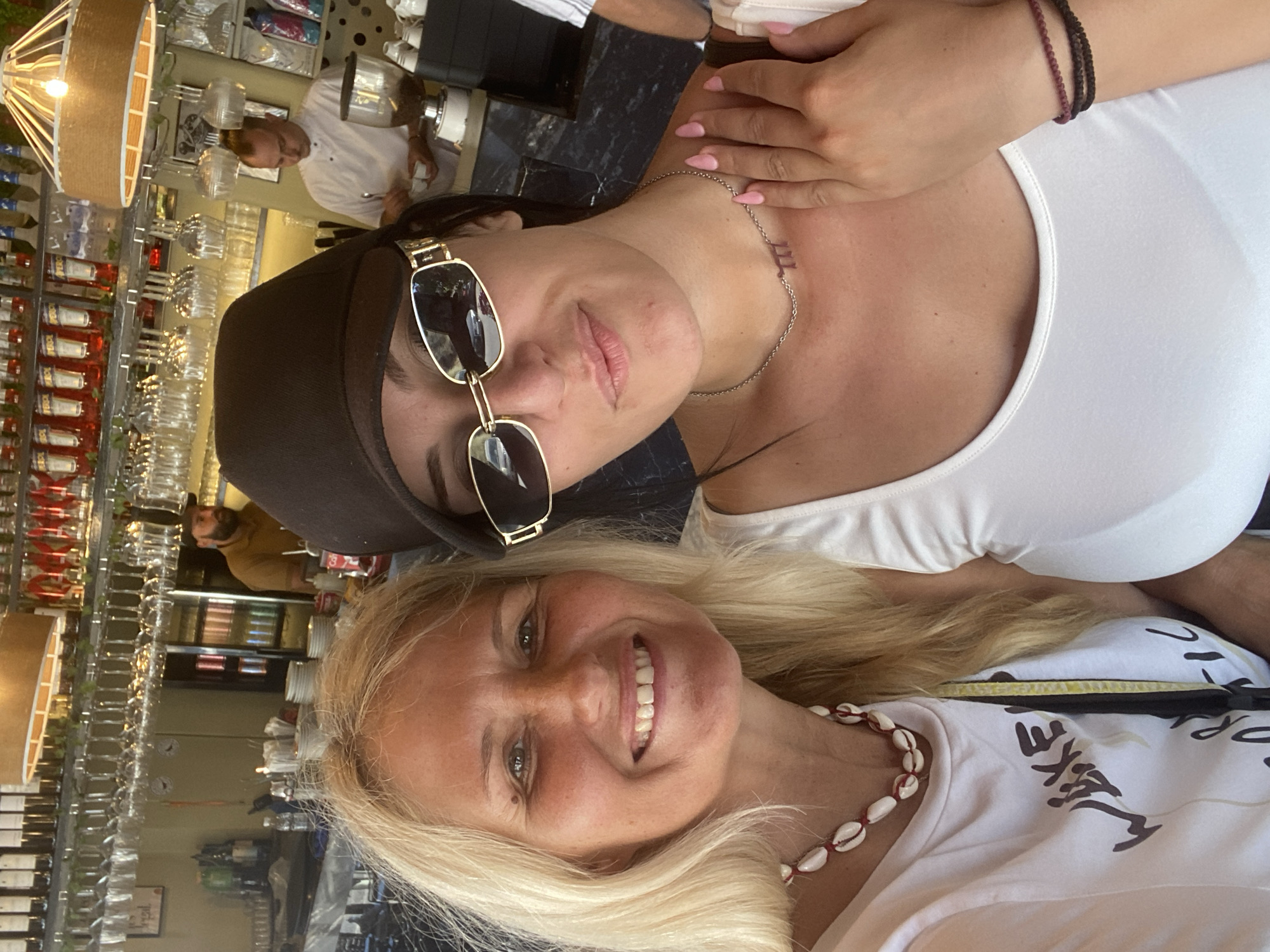 Two women are taking a selfie in what appears to be a bar or restaurant. The woman on the left has blonde hair and is wearing a white t-shirt and a shell necklace. The woman on the right has dark hair and is wearing a white top and a dark cap, with sunglasses on her nose. Behind them, shelves are stocked with bottles and glasses. A man in a white chef's uniform is visible in the background working at a counter. The location is identified as Ia Municipal Unit, Greece.