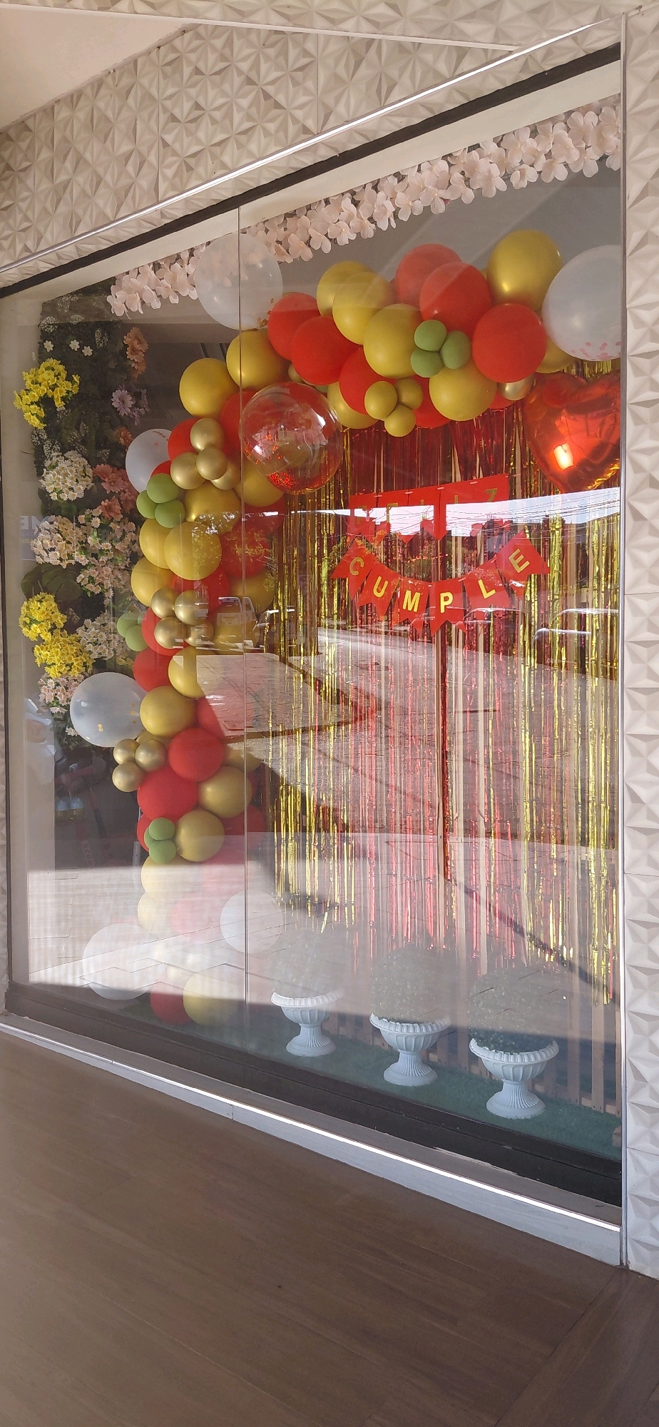 A storefront window display in Encarnación, Paraguay, features a vibrant balloon arch and festive decorations. The balloons are a mix of red, yellow, gold, and white, with some green accents. A banner reading "FELIZ CUMPLE" (Happy Birthday in Spanish) is hung in front of a backdrop of gold and red fringe curtains. In the foreground, three white decorative urns with textured tops are visible. The window reflects the street outside, showing glimpses of cars and buildings. The overall impression is a lively celebration setup, likely for a birthday event.