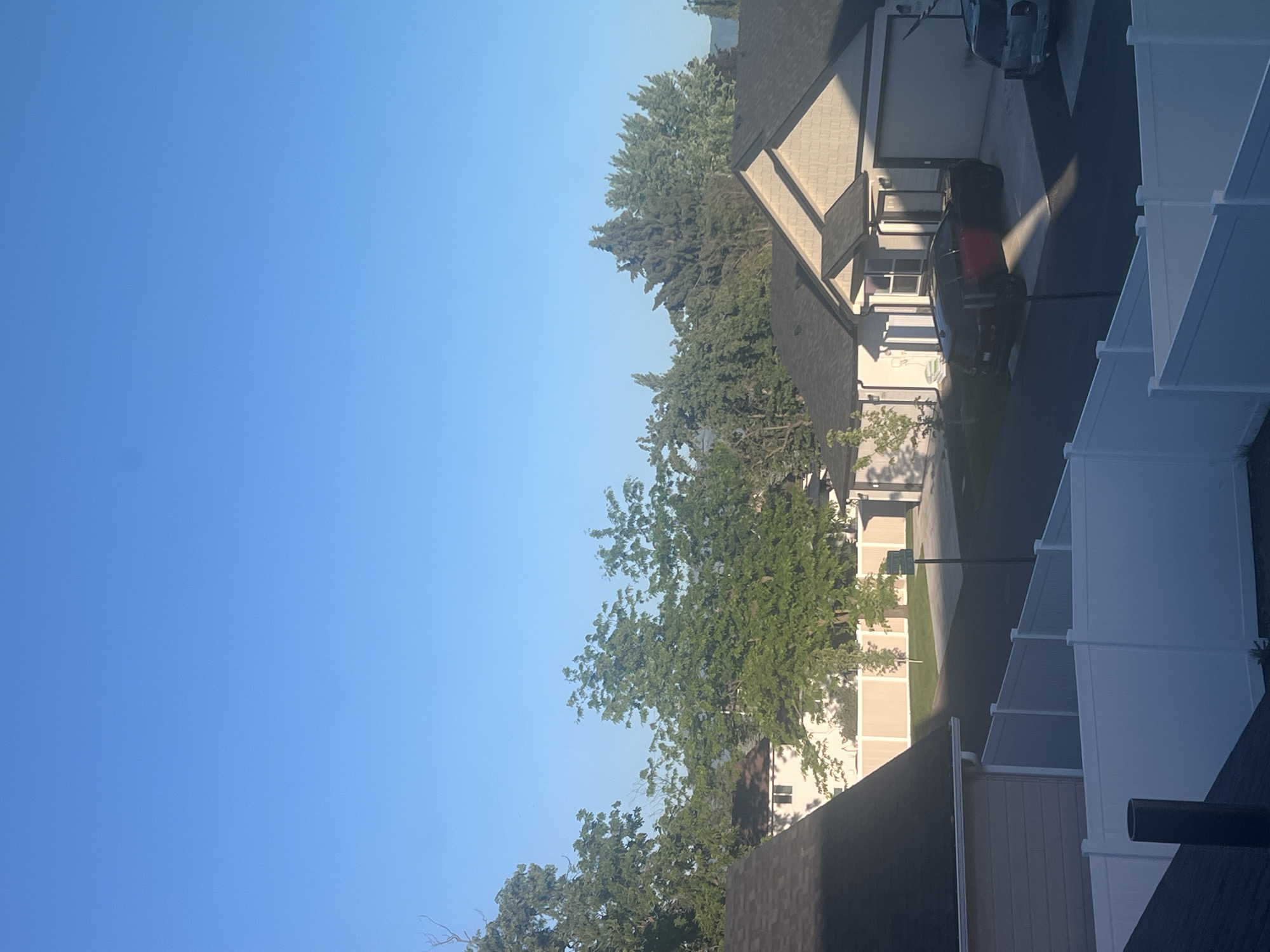 The image shows a suburban street in Spokane Valley, USA, under a clear blue sky. The foreground features a white vinyl fence. Behind the fence, a dark asphalt road curves through a residential area. Several houses with dark roofs are visible, some with attached garages. A dark-colored SUV is parked in a driveway, and another vehicle is partially visible on the right side of the frame. Mature trees with lush green foliage line the street and surround the properties. The lighting suggests it is morning or late afternoon, with long shadows cast across the scene.