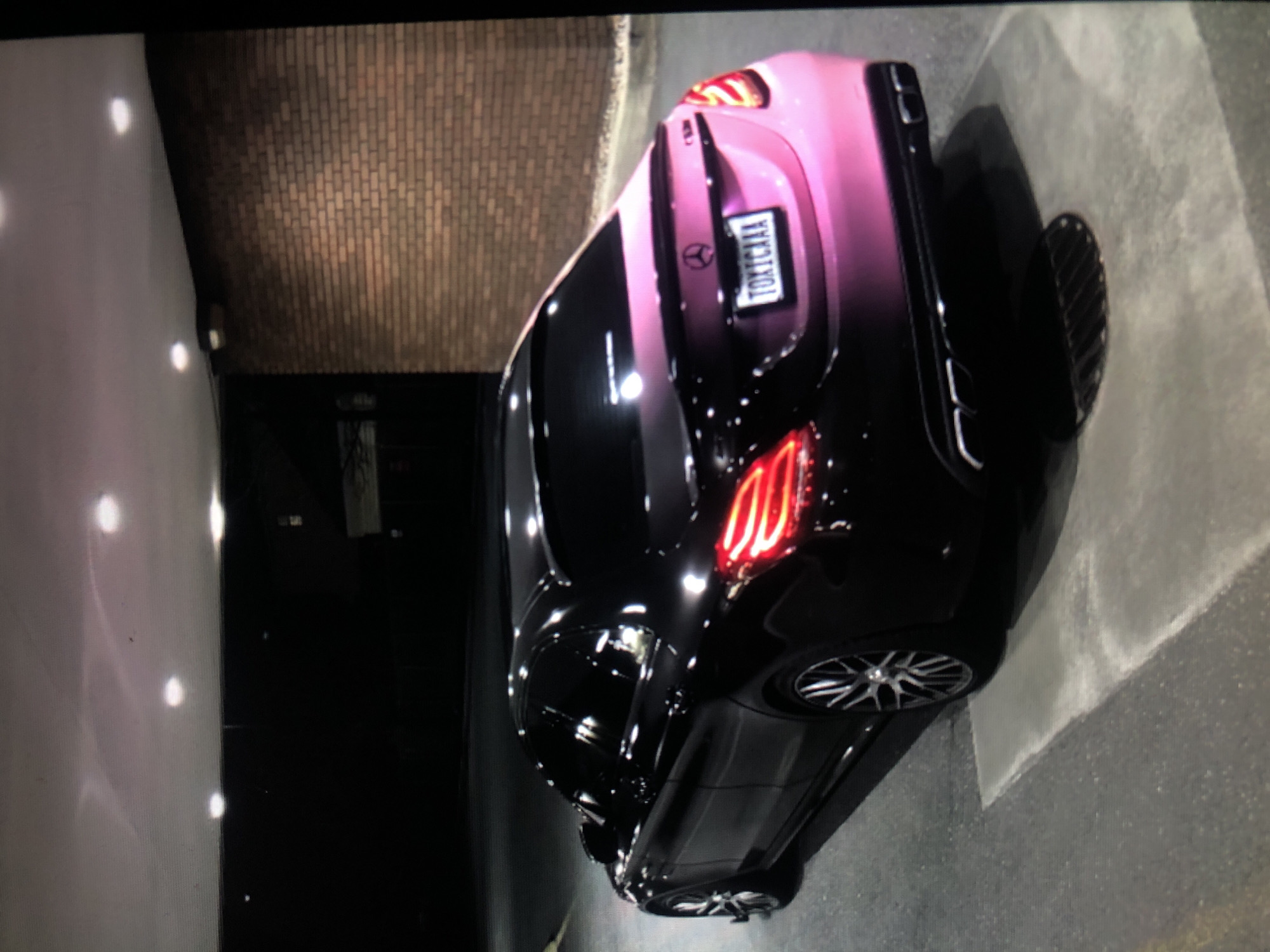 The image shows a black and pink Mercedes-Benz parked in what appears to be an alley or a confined parking area. The car's rear is visible, featuring the license plate "TOKICAAA." The location appears to be an urban environment. Based on the presence of the license plate and the car's make, this location is not Lhokseumawe, Indonesia. The environment includes a brick wall, concrete ground, and a white wall illuminated with several lights.