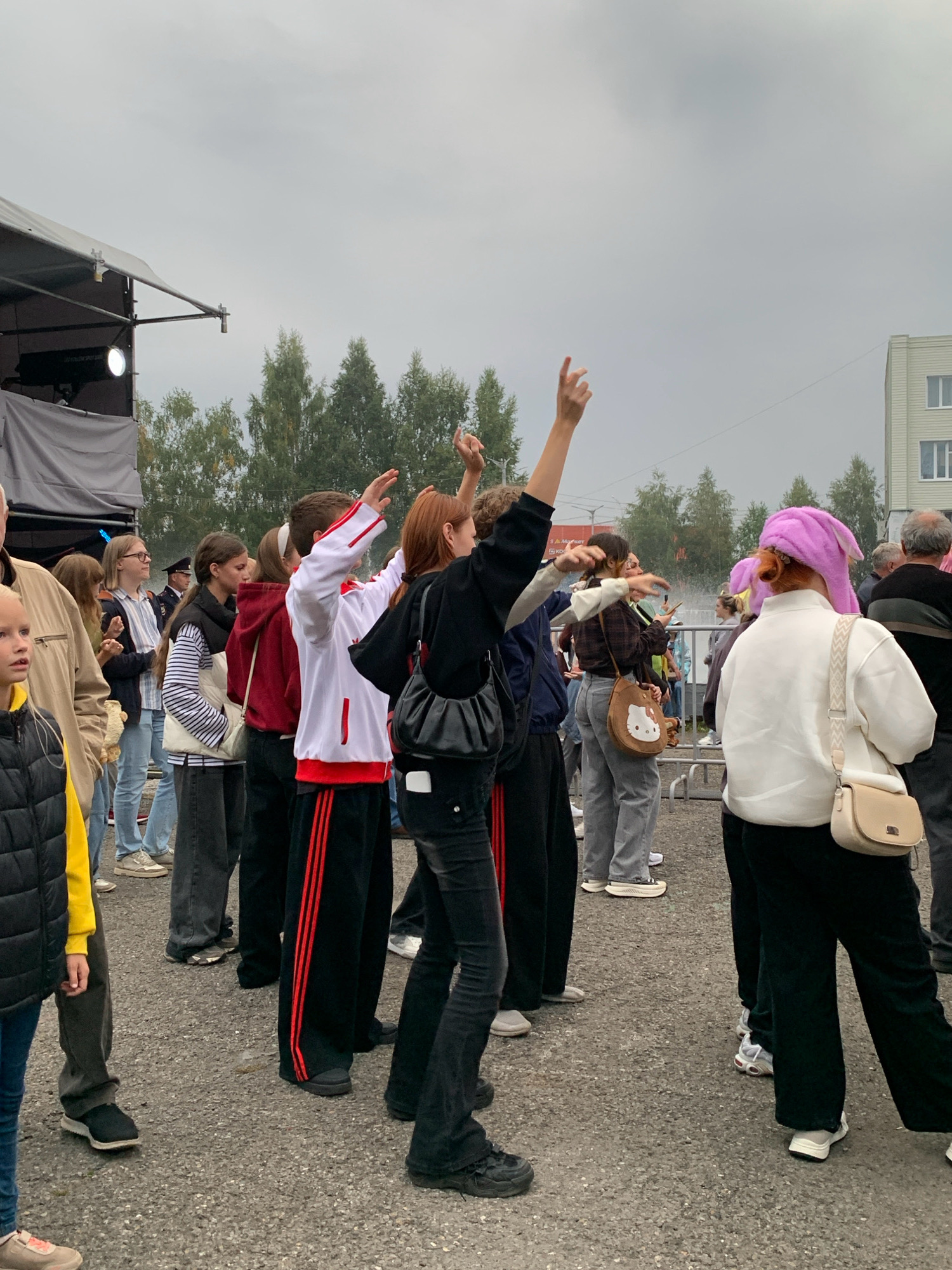 In Prokopyevsk, Russia, a group of young people stand outdoors, their backs to the camera, observing an event. The sky is overcast, and the ground is paved with asphalt. In the foreground, a young girl with blonde hair wearing a black vest over a yellow shirt is partially visible. To her left, a man in a beige jacket and black pants stands. The central focus is a group of teenagers; one in the foreground wears a black hoodie and black flared pants, her arms raised. Next to her, another teenager in a white hoodie with red accents and black pants with red stripes also has their arms raised. Further back, a young woman with red hair wears a pink bunny hat and a white jacket, with a beige purse slung across her body. Other individuals are visible in the background, some with their arms raised. Behind the crowd, trees and a light-colored building are visible. A stage structure with a gray tarp and a spotlight is present on the left side of the image.
