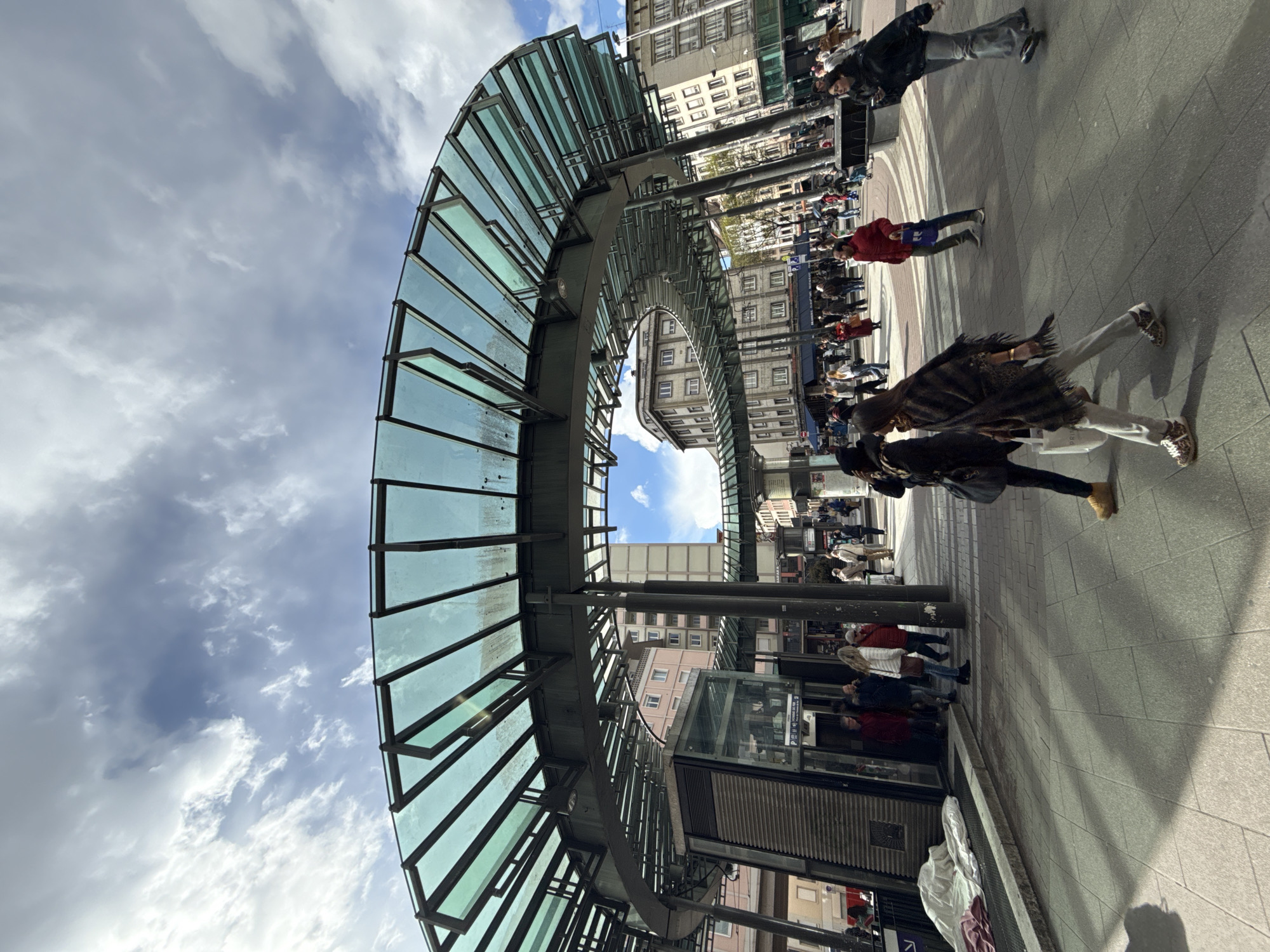 The image displays an urban plaza scene at Place d'Austerlitz in Strasbourg, France, centered around a distinctive, curved glass and metal canopy structure.
The primary architectural feature is a large, semi-circular canopy constructed from a dark metal framework supporting numerous green-tinted glass panels. This structure is supported by multiple thick, dark cylindrical pillars. Beneath a segment of the canopy, a multi-faceted dark-ribbed metal and glass structure forms an entrance, likely to an underground transit station, indicated by visible signage including a white 'P' symbol and French text. Adjacent to this entrance, a rolled item, possibly a sleeping bag or blankets, covered by a white sheet, rests on a raised platform.
The ground consists of light-colored square and rectangular paving tiles, showing elongated shadows cast from the left.
Multiple pedestrians populate the scene. In the immediate foreground, two women are walking from right-to-left across the pavement. The woman in front wears a dark coat and light-colored pants, holding a white shopping bag. The woman behind her wears a patterned brown and black poncho, light pants, and leopard-print shoes, also carrying a white shopping bag. Further back, a man in a red jacket and blue jeans walks from left-to-right, carrying a purple bag. To the far right, a woman in a dark jacket and light flared jeans holds what appears to be a mobile device. A group of individuals is gathered near the station entrance, and numerous additional people are visible dispersed across the paved square and along the surrounding streets.
The background features multi-story buildings typical of a European city, exhibiting various architectural styles with facades of beige and light colors and numerous windows. The sky overhead is partly cloudy, with patches of blue sky visible between scattered white clouds, particularly through the open central area of the canopy structure.