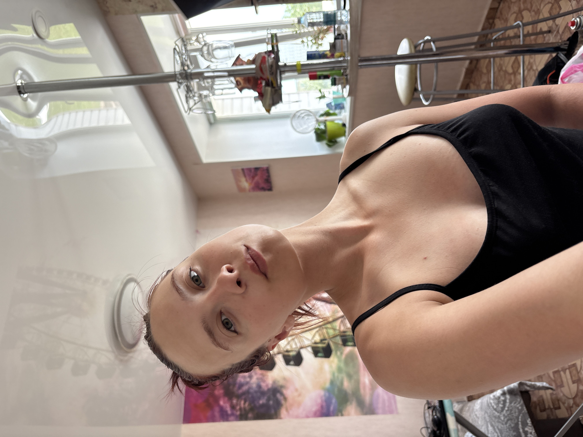 The image is a selfie taken from a low angle by a young woman with fair skin and light eyes. She is wearing a black top with thin straps. Her hair is reddish-brown and appears slightly damp.
The background appears to be an interior setting, possibly an apartment. A window with a view of greenery outside is visible. Above the window is a shelf with various items. Below the woman, there's a decorative wall hanging with abstract colors. A stainless steel bar and bar stools are visible further in the background. A circular object, possibly a light fixture, is seen behind and to the left of her head.
The location might be in or near Прикамский, Russia, if that is relevant to identifying objects or features in the image.