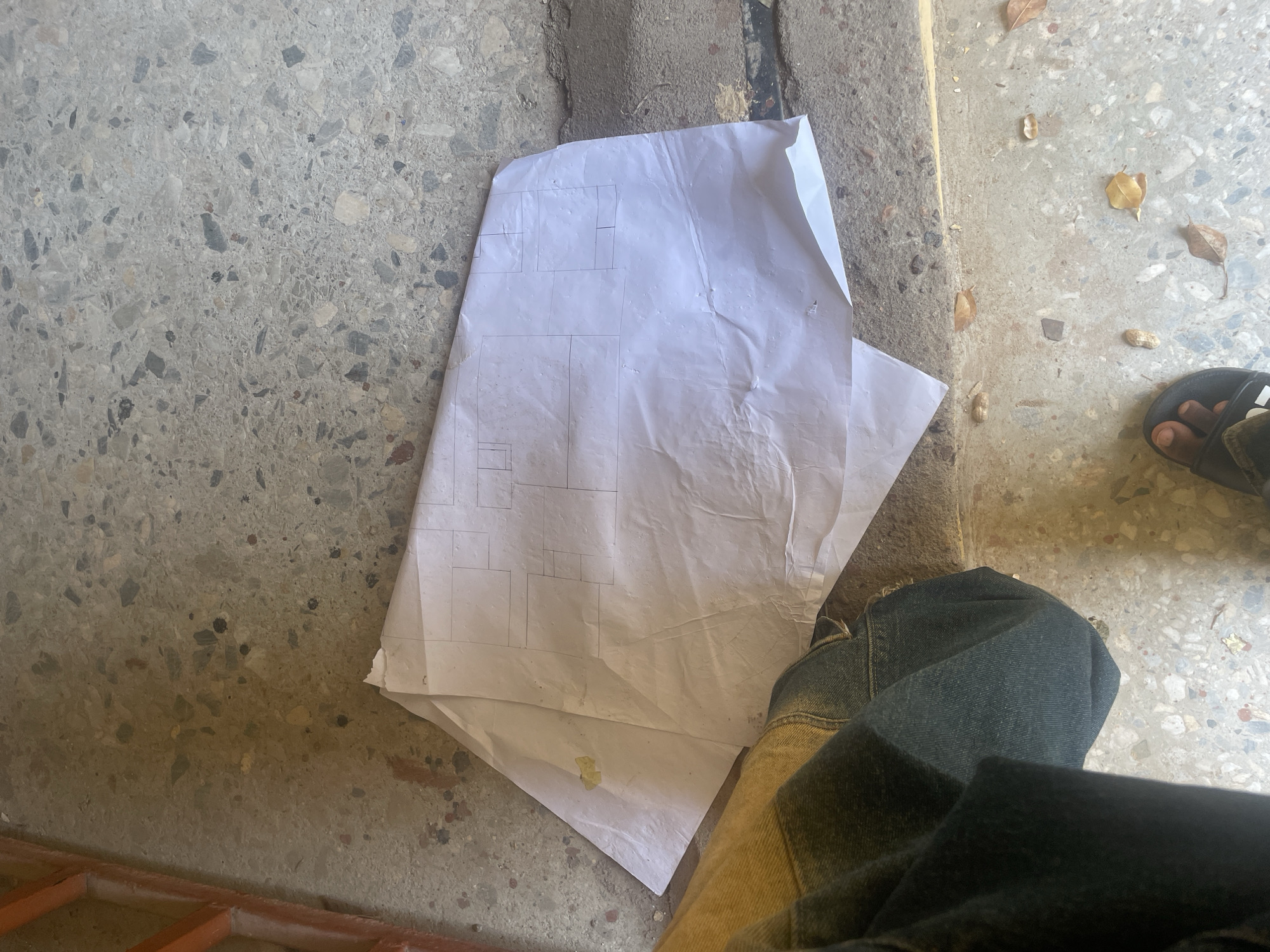 An outdoor scene in Lokoja, Nigeria, features a crumpled white paper with hand-drawn lines, resembling a floor plan or blueprint, lying on a textured concrete surface. The paper is partially obscured by the leg of a person wearing denim jeans and a yellow and brown patterned garment. The concrete floor is speckled with various shades of gray, white, and black aggregate, with some areas appearing worn and stained. Dry leaves and small debris are scattered on the ground. In the lower left portion of the image, a sandal is visible on a foot. To the left of the paper, a portion of an orange metal grate is visible. The lighting suggests it is daytime.