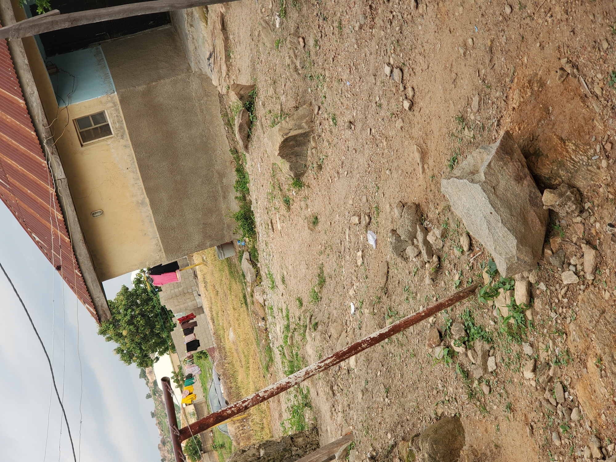The image depicts an outdoor scene, possibly a rural or semi-urban area, with a focus on a building and the surrounding unpaved ground. A building with a rusted corrugated metal roof is visible on the left, partially obscuring a section of the building's wall which is painted yellow with a light blue accent near the top. A small window with a grid pattern is present on the yellow wall. Below the window, a light fixture is attached to the wall.
In front of the building, clothes are hanging on a clothesline. The clothes are of various colors, including pink, black, red, brown, and yellow. A green tree with dense foliage stands to the left of the clothesline. The ground in the foreground is dry and sandy, scattered with numerous rocks and stones of varying sizes. Some small green plants are growing between the rocks and in patches of soil. A rusty metal pole, likely part of a fence or support structure, extends diagonally across the lower part of the frame. In the background, there appears to be more construction, possibly brick buildings, and a landscape with rolling hills and scattered rocks. The sky is overcast. The scene suggests a daytime setting. No people are clearly visible.